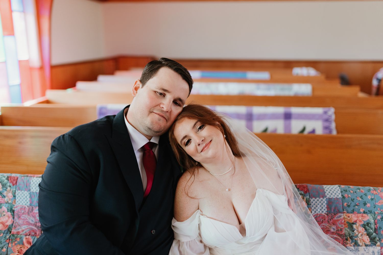 A couple in formal wedding attire share a romantic moment while sitting in wooden church pews.