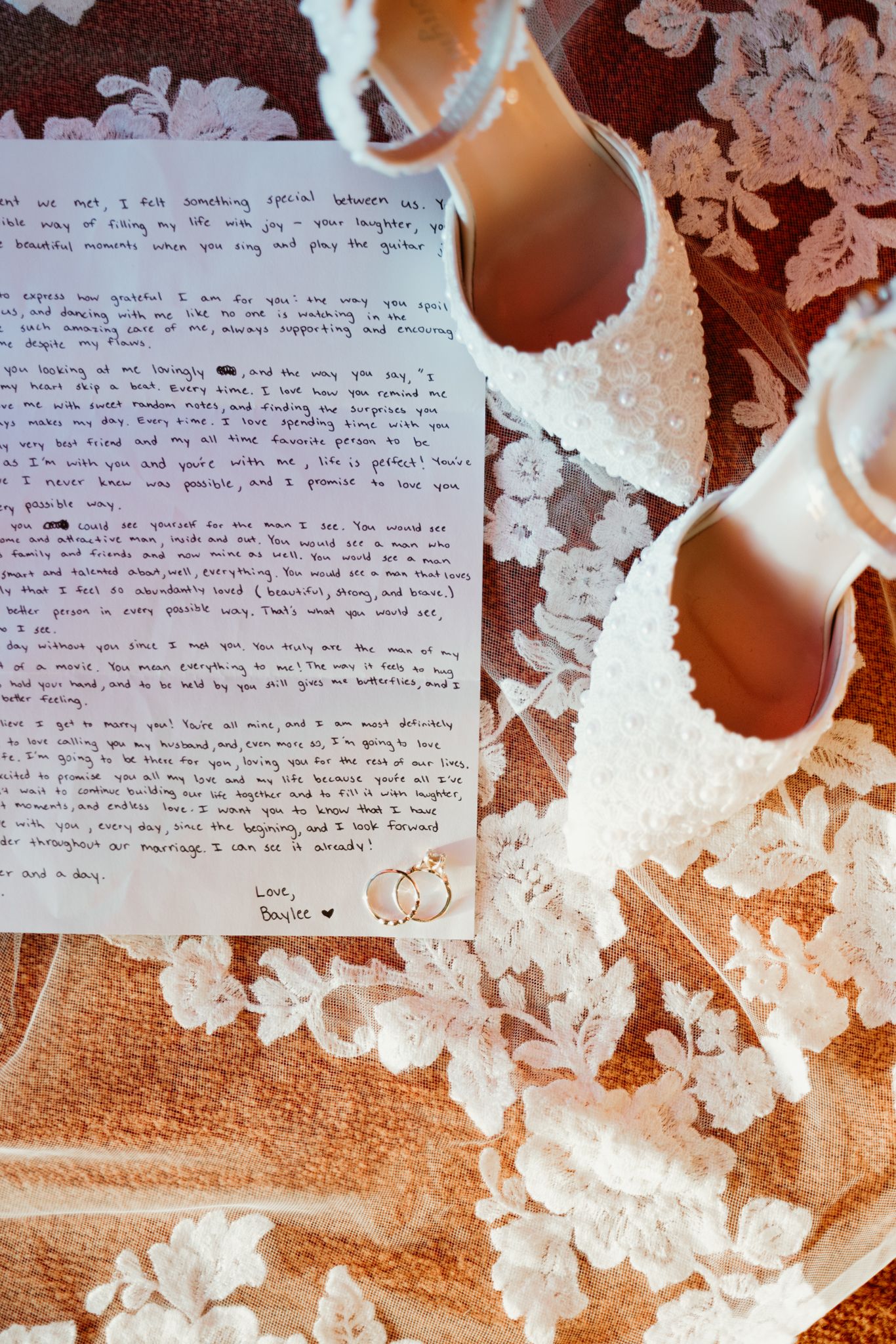 A handwritten love letter alongside delicate white lace wedding shoes on a floral patterned background.