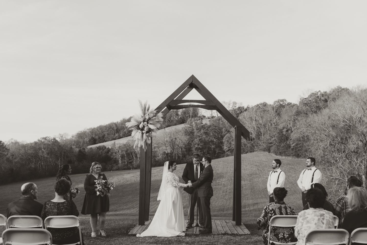 A black and white photograph of an outdoor wedding ceremony under a wooden arch with guests seated in chairs.