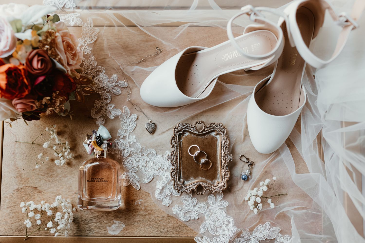 Wedding accessories including elegant white heels, perfume bottle, delicate flowers and rings displayed on a vintage table.