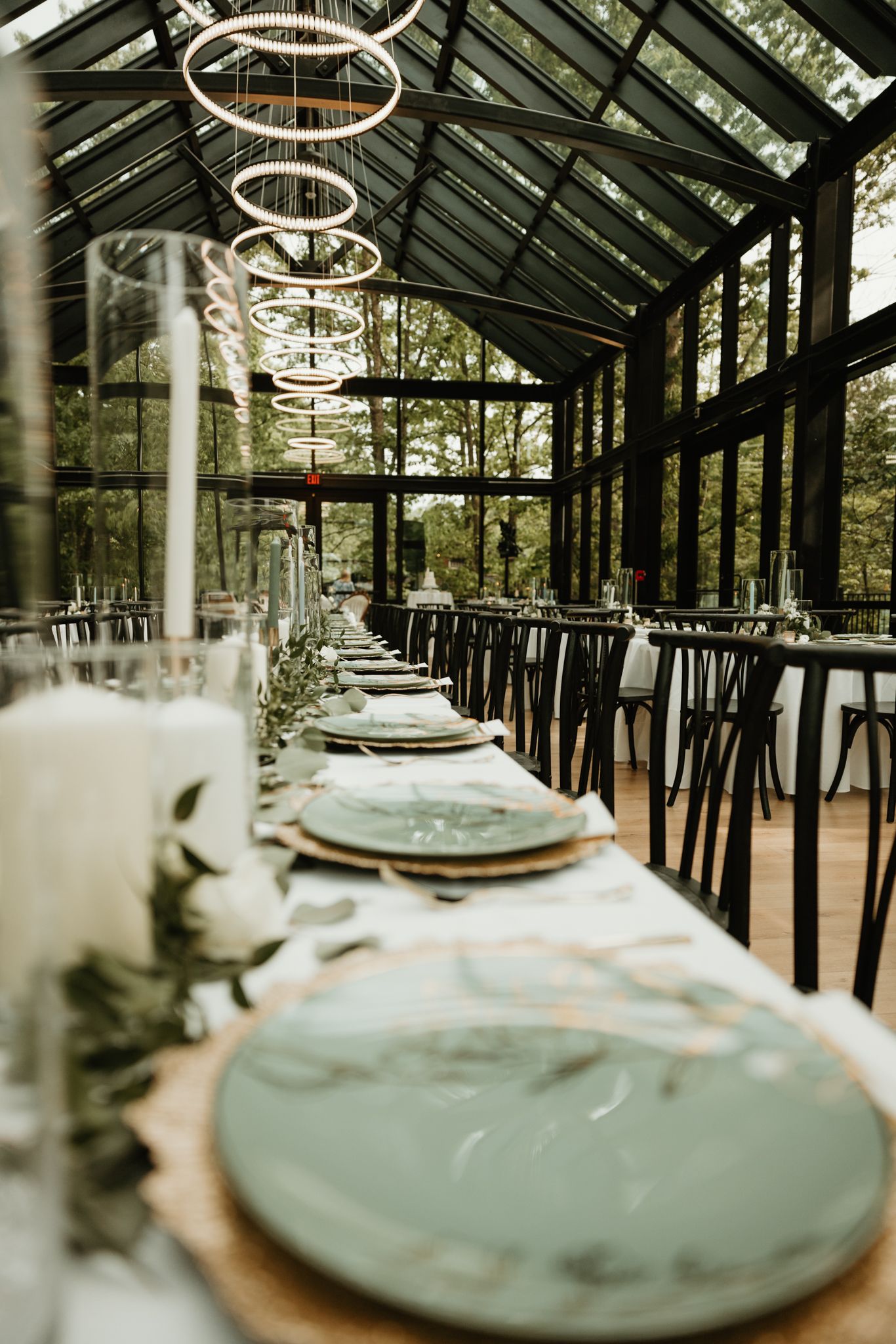 Elegant greenhouse dining setup with sage green plates and modern decor featuring circular pendant lights and glass walls.