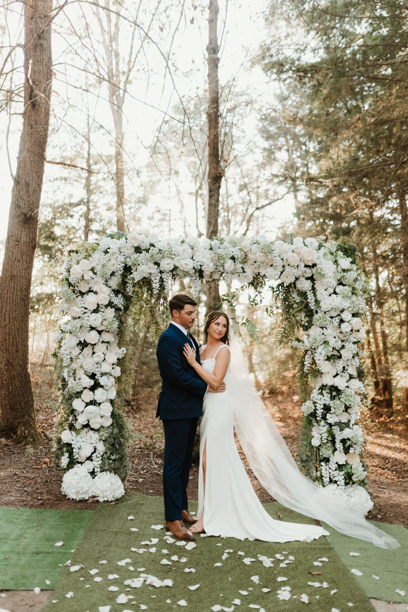 White floral wedding arch in a wooded outdoor setting with scattered rose petals on green grass.