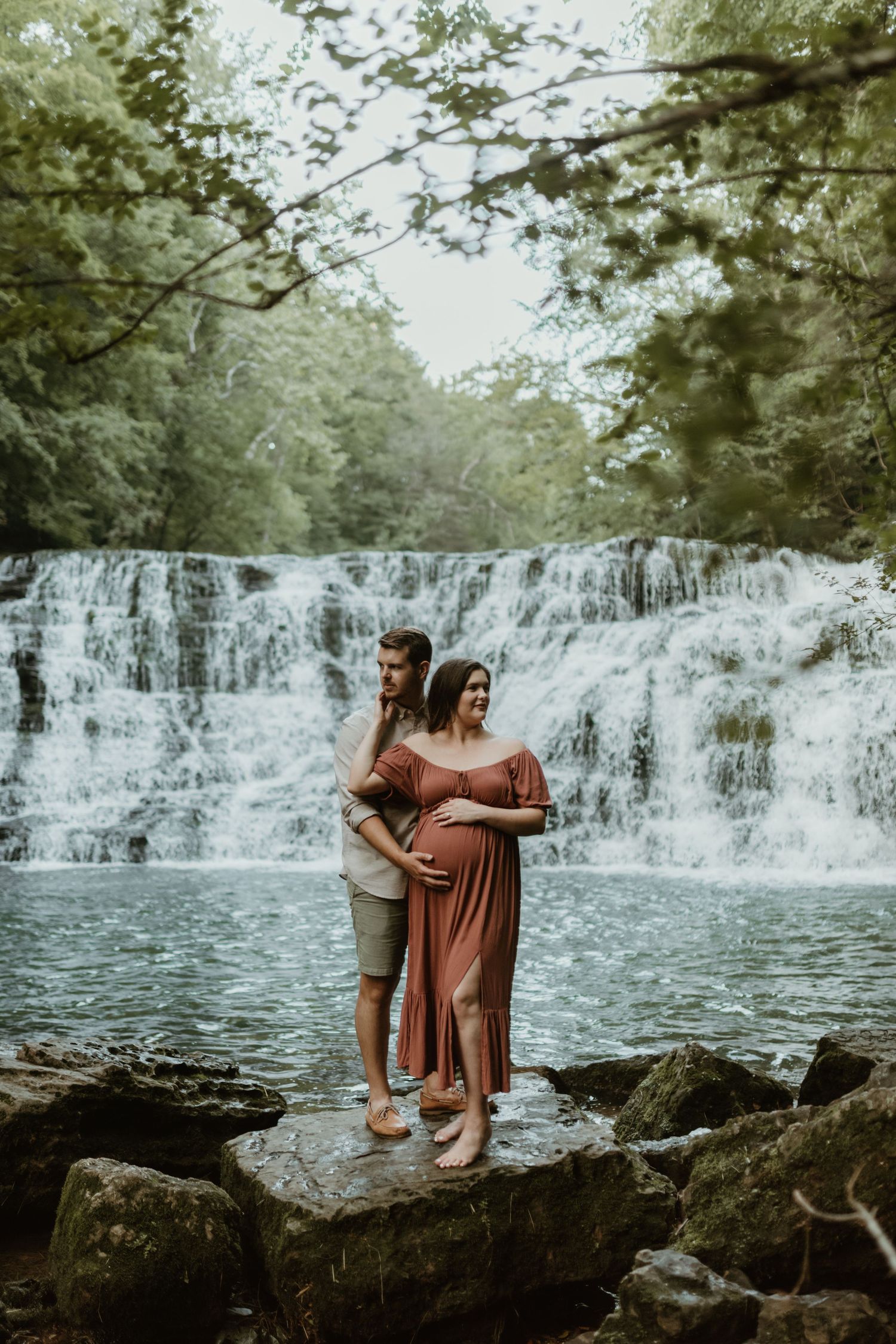 Couple stands together on rocks overlooking a cascading waterfall in a romantic outdoor setting.