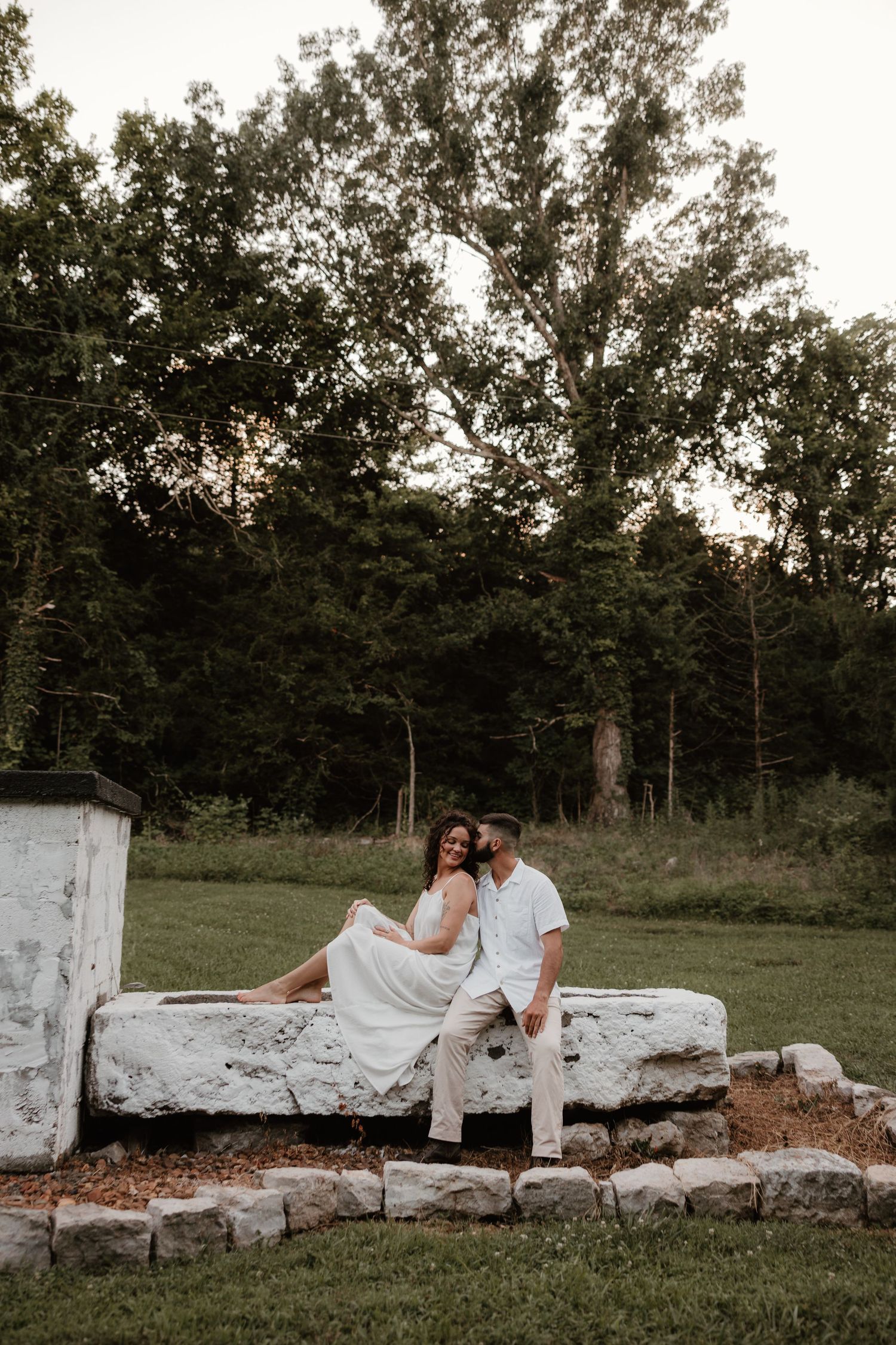A couple in wedding attire sits intimately on white stone blocks outdoors at dusk.