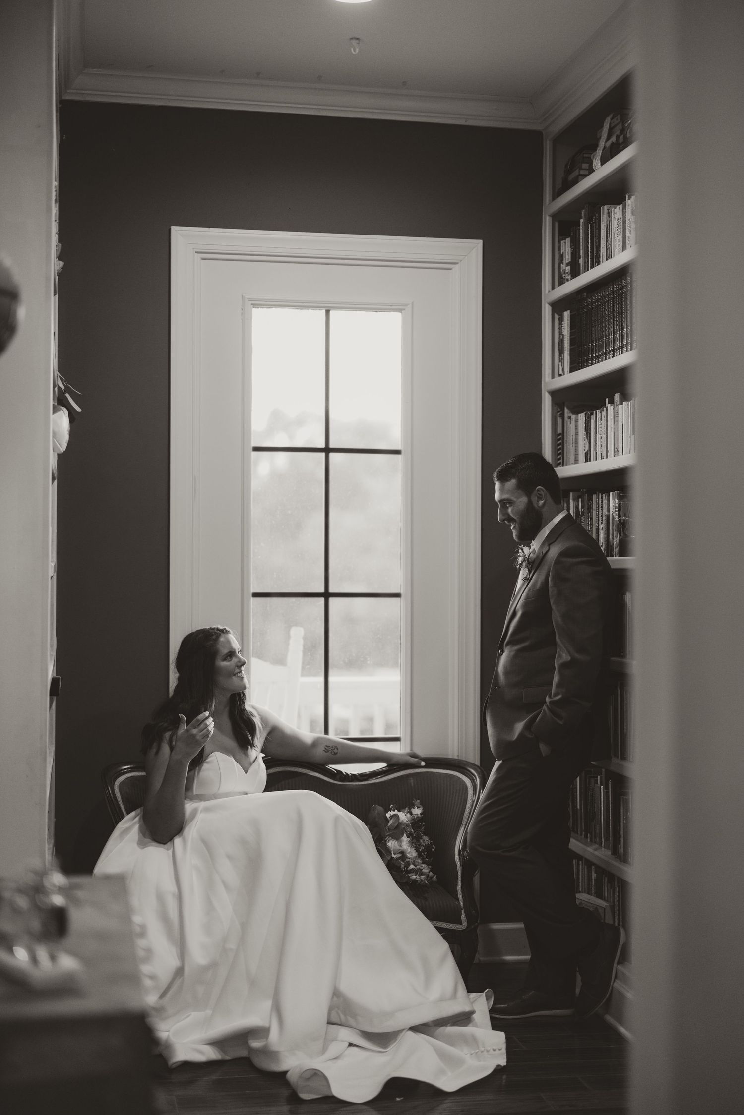 Black and white wedding photo of bride in gown seated by window as groom leans against bookshelf.