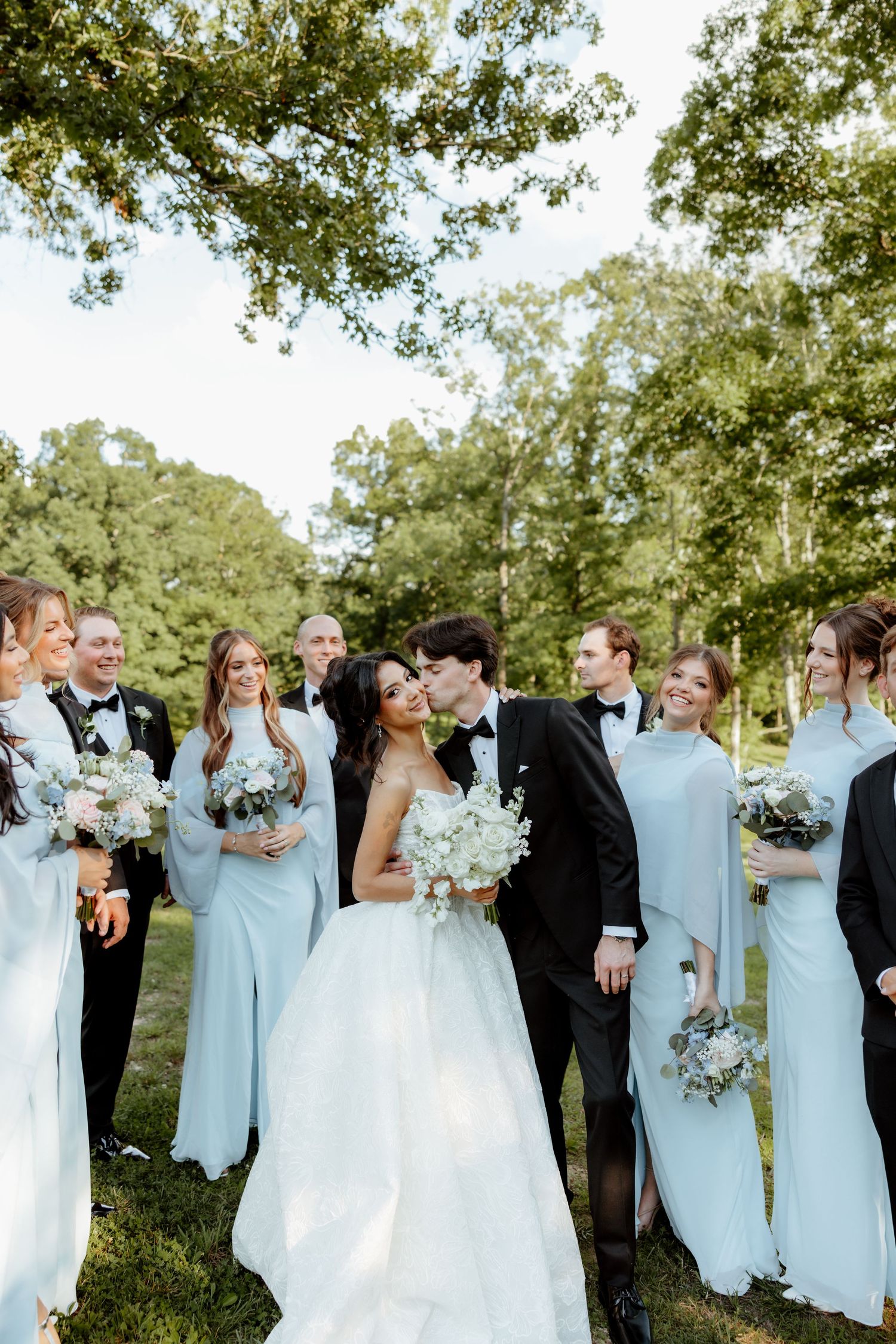 Wedding party in light blue gowns and black tuxedos surrounds kissing bride and groom outdoors.