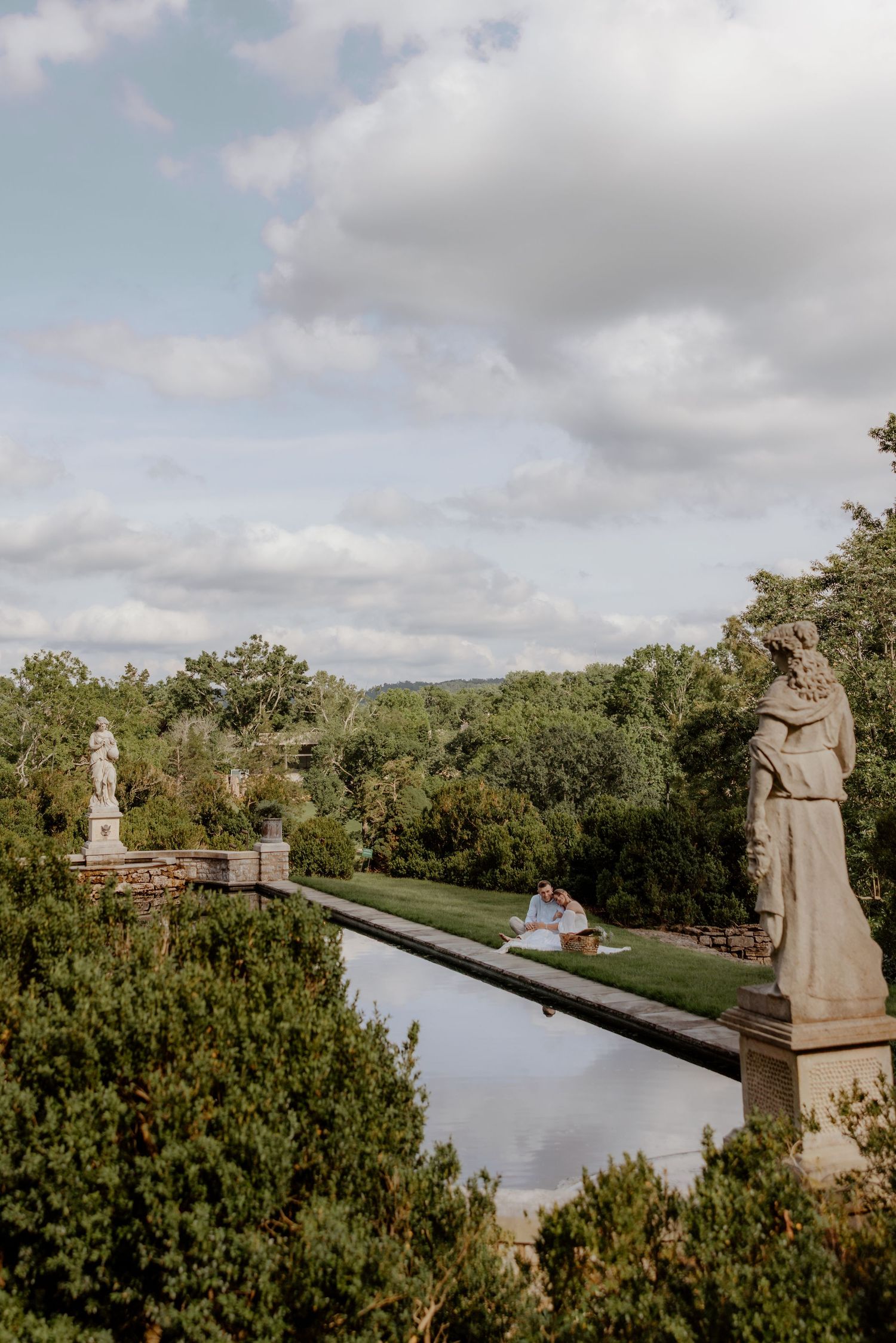 Couple sitting beside a formal reflecting pool flanked by classical stone statues in a lush garden landscape.