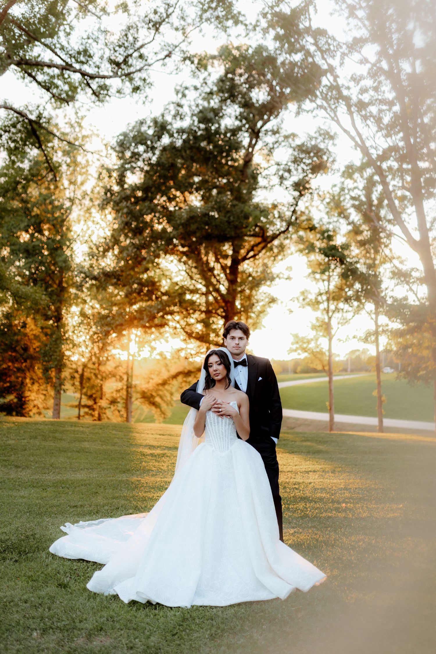 Bride in white ball gown and groom in black tuxedo embrace at golden hour in a lush green park setting.