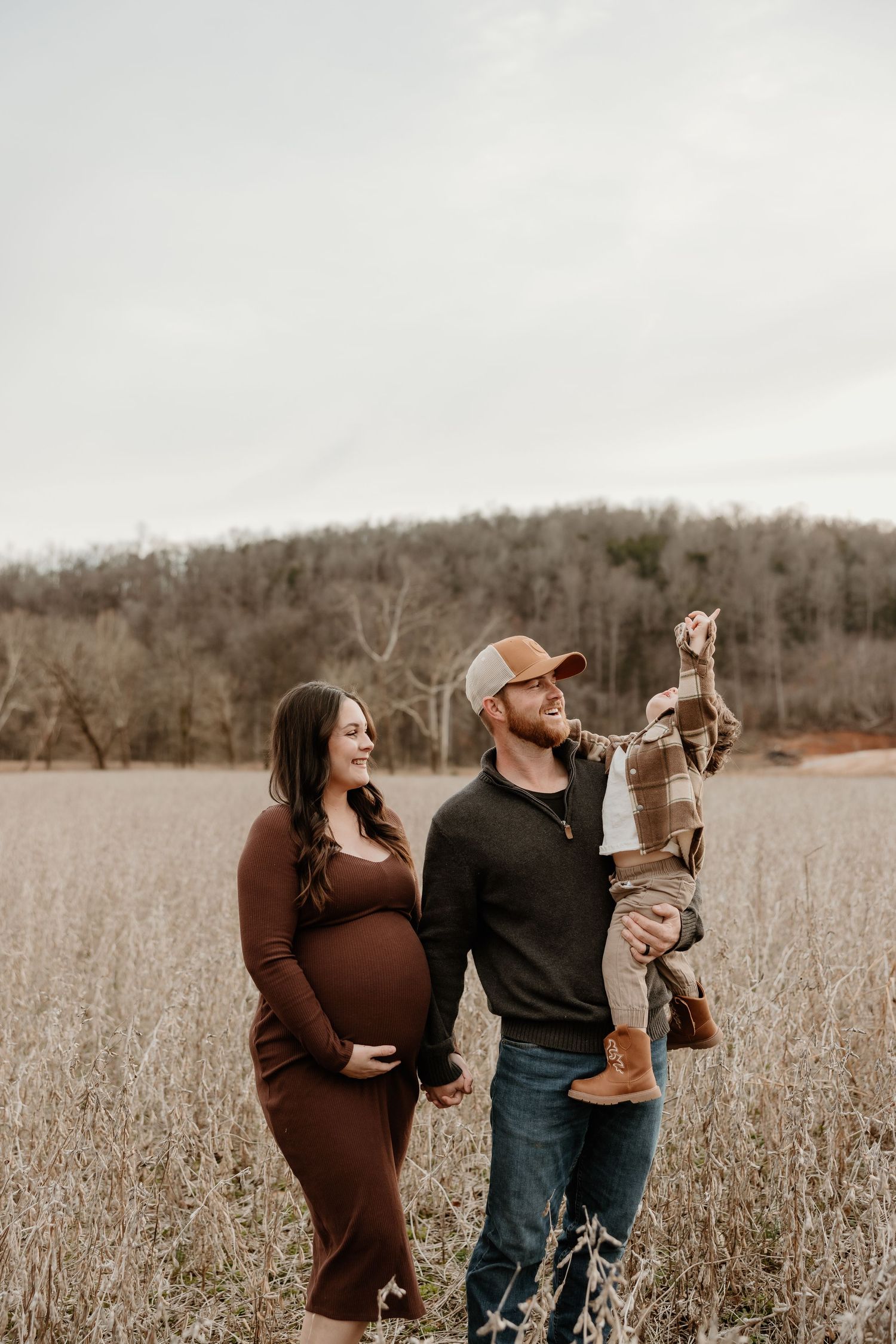 Pregnant woman in brown dress with husband holding toddler in autumn field maternity photo session.