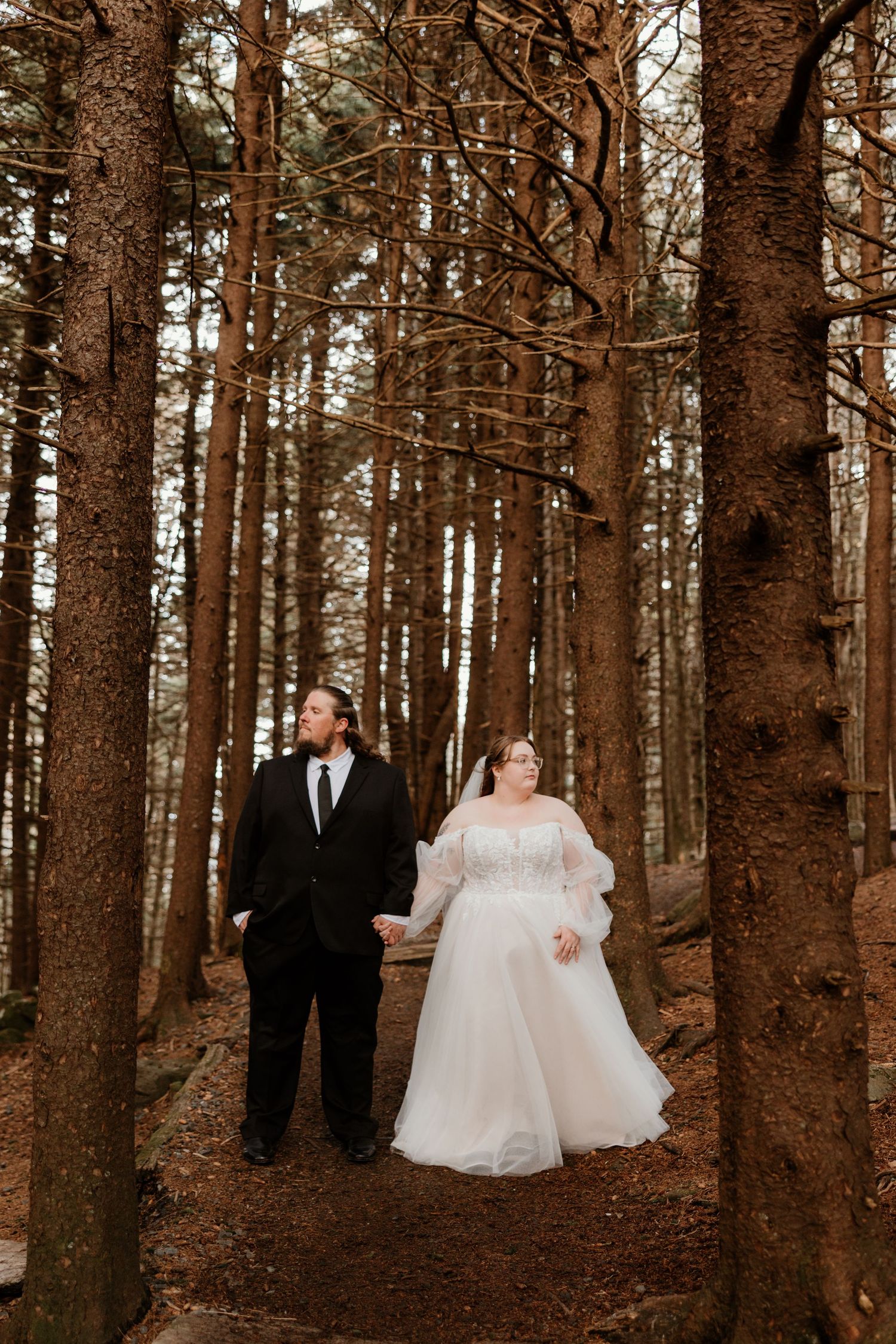 Bride and groom hold hands walking through tall pine forest in elegant black suit and white tulle wedding gown.