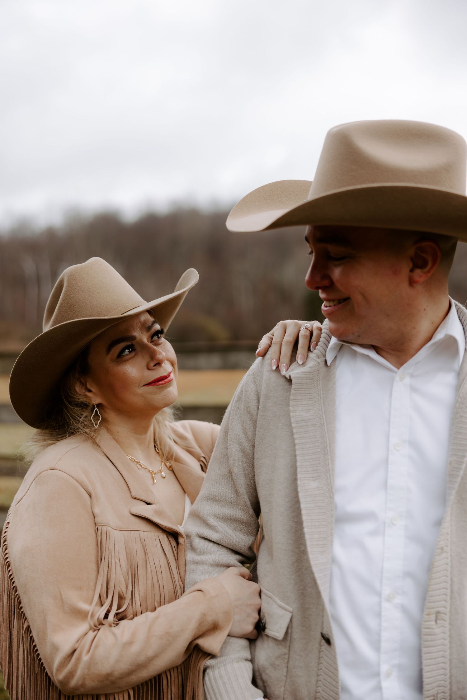 Couple in matching tan cowboy hats embrace outdoors, woman in fringe jacket looks up smiling at man in white shirt.