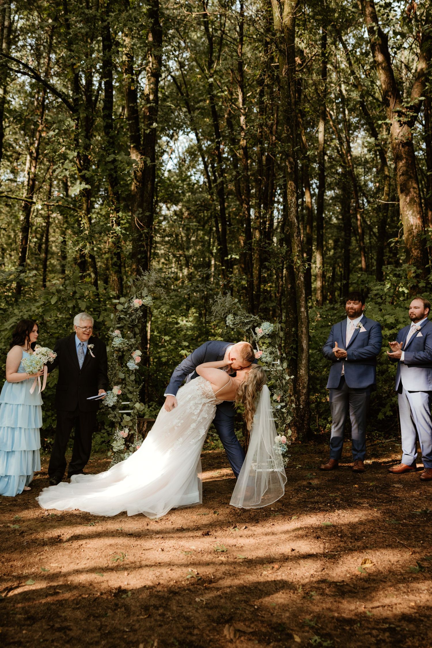 A wedding ceremony taking place in a sunlit forest with guests gathered on a dirt path surrounded by tall trees.