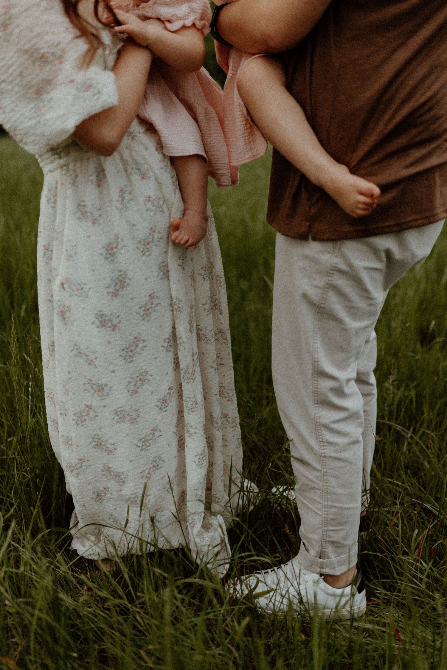 Two parents holding a baby in a green field, wearing floral dress and brown shirt.