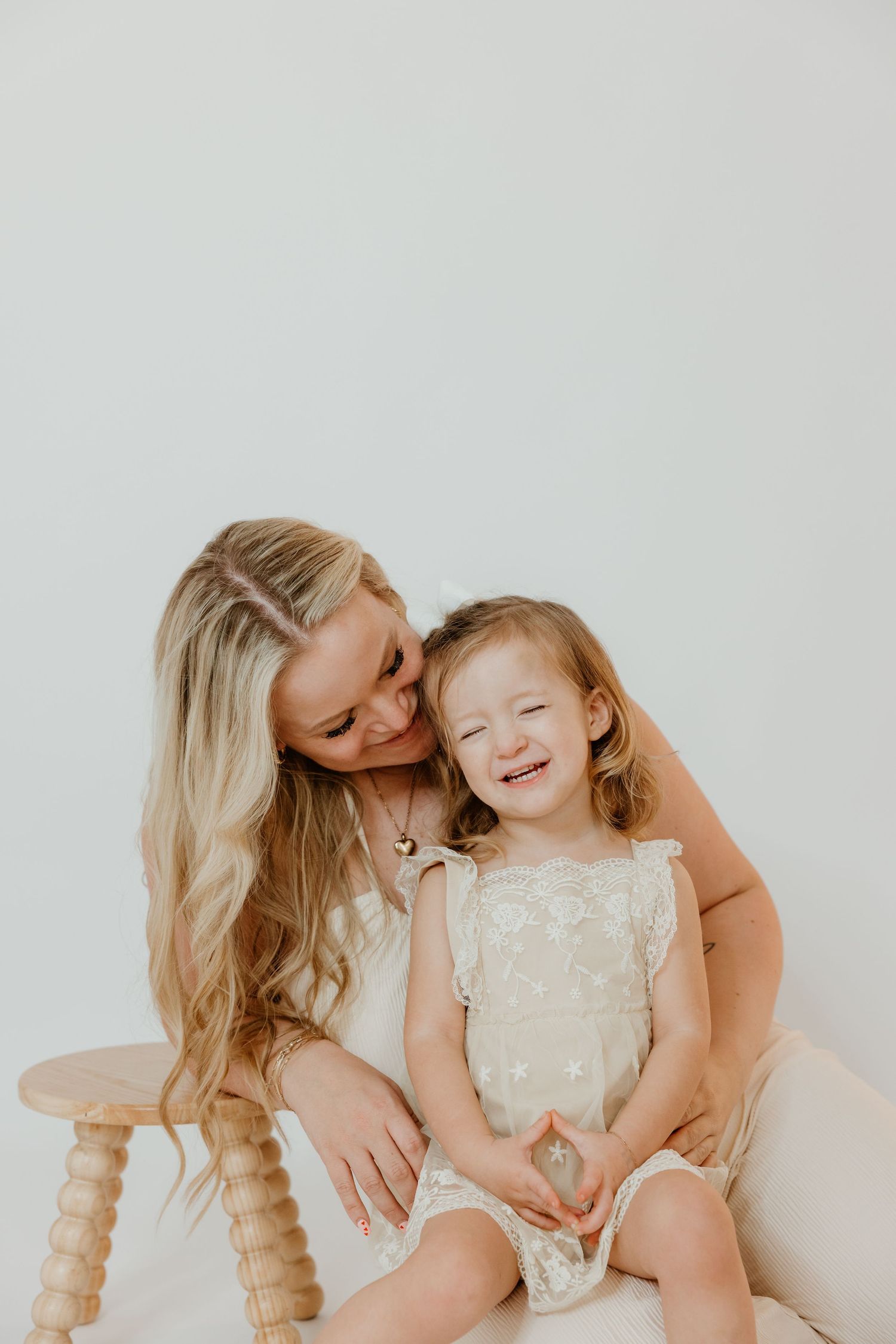 A smiling blonde mother kisses her laughing toddler daughter, both wearing cream dresses, seated by a wooden stool.