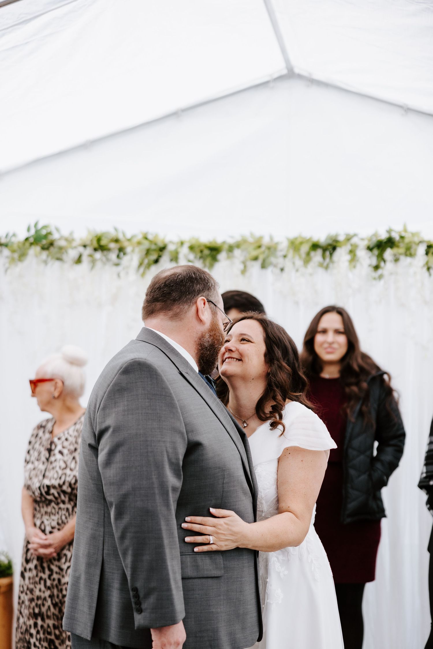Bride and groom share a joyful moment at their wedding ceremony under a white tent adorned with greenery.
