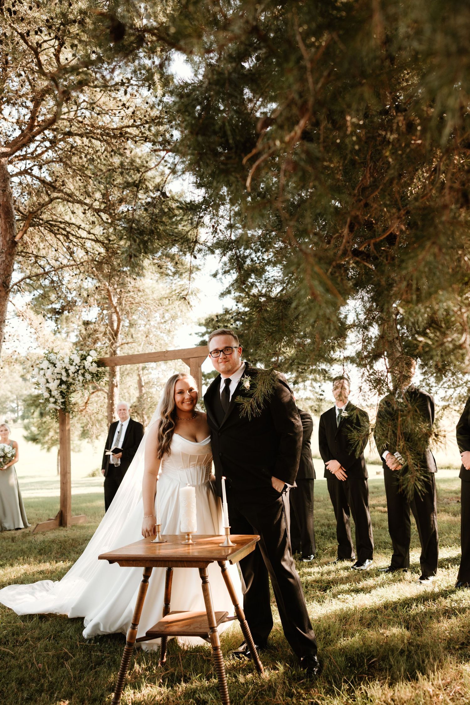 A wedding ceremony takes place outdoors with attendants standing in a line under tall trees.