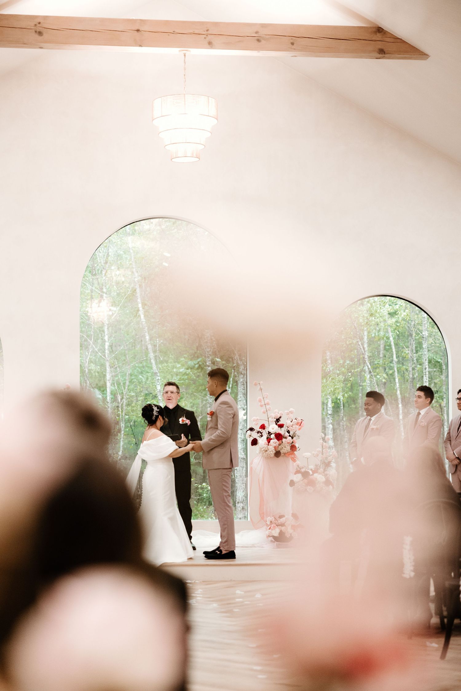 An intimate wedding ceremony takes place in a bright white chapel with natural light streaming through arched windows.