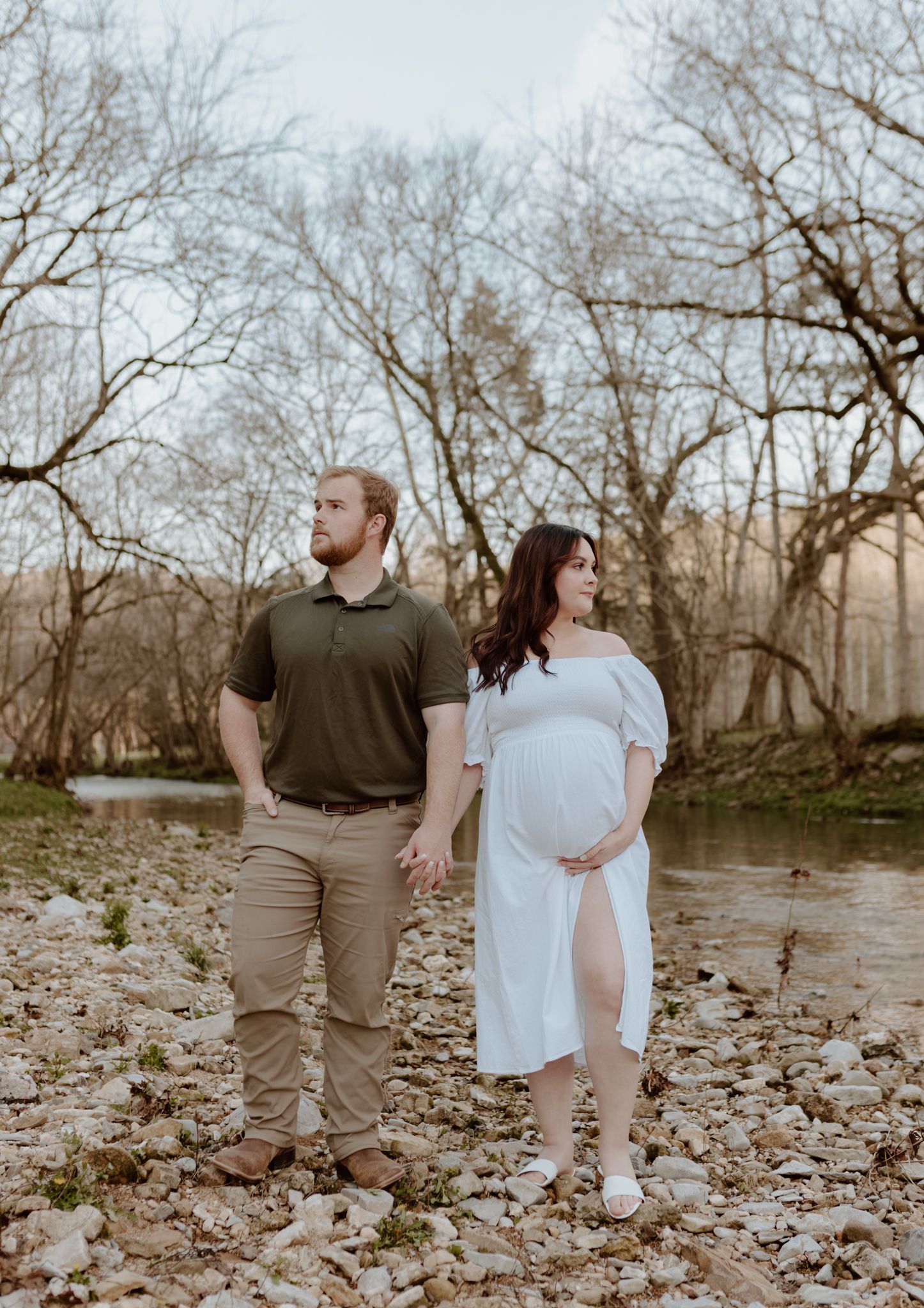 A couple takes a romantic stroll along a rocky riverbank surrounded by bare winter trees.