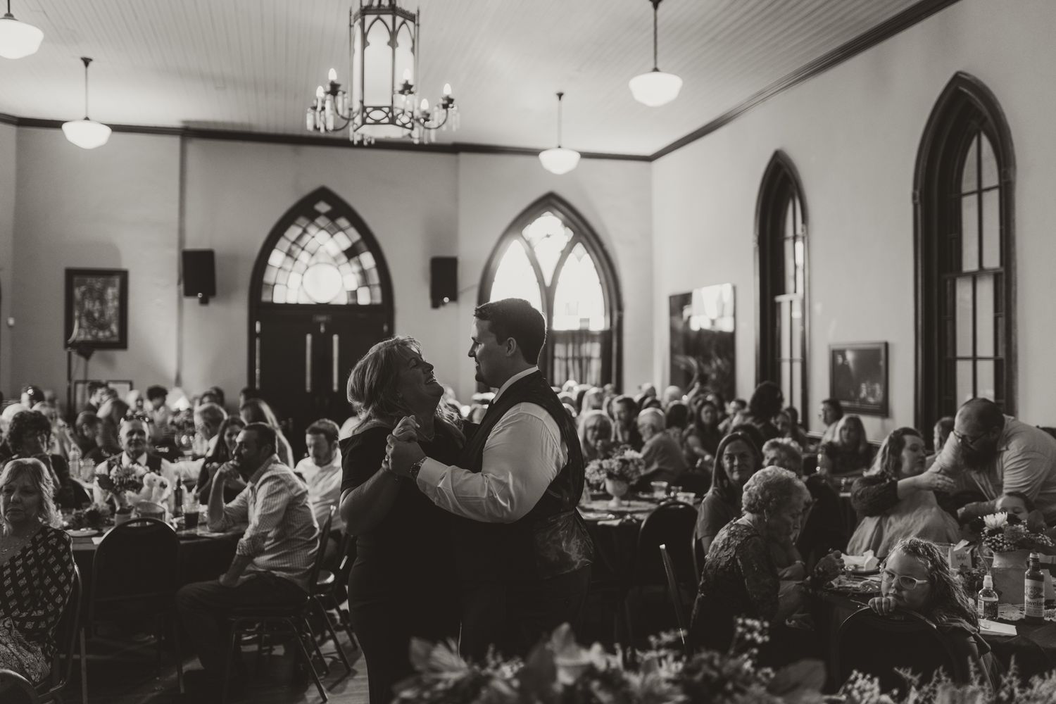 Black and white photo of a wedding reception scene in an elegant room with arched windows and a chandelier.