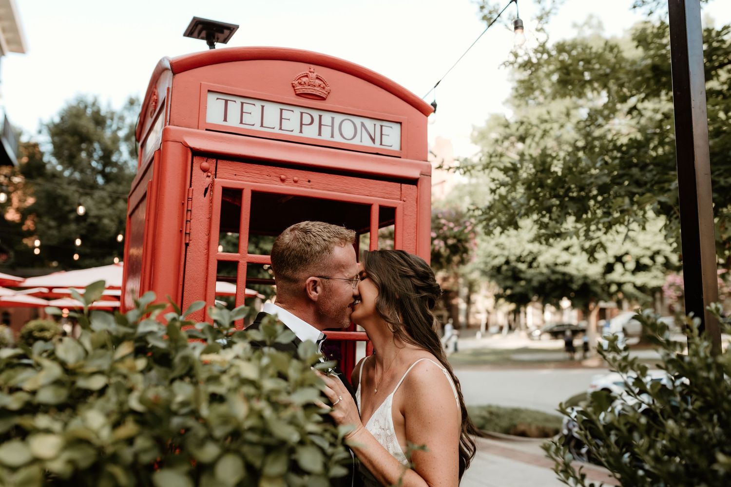 Two people standing next to a vintage red British telephone booth share a romantic moment in a garden setting.
