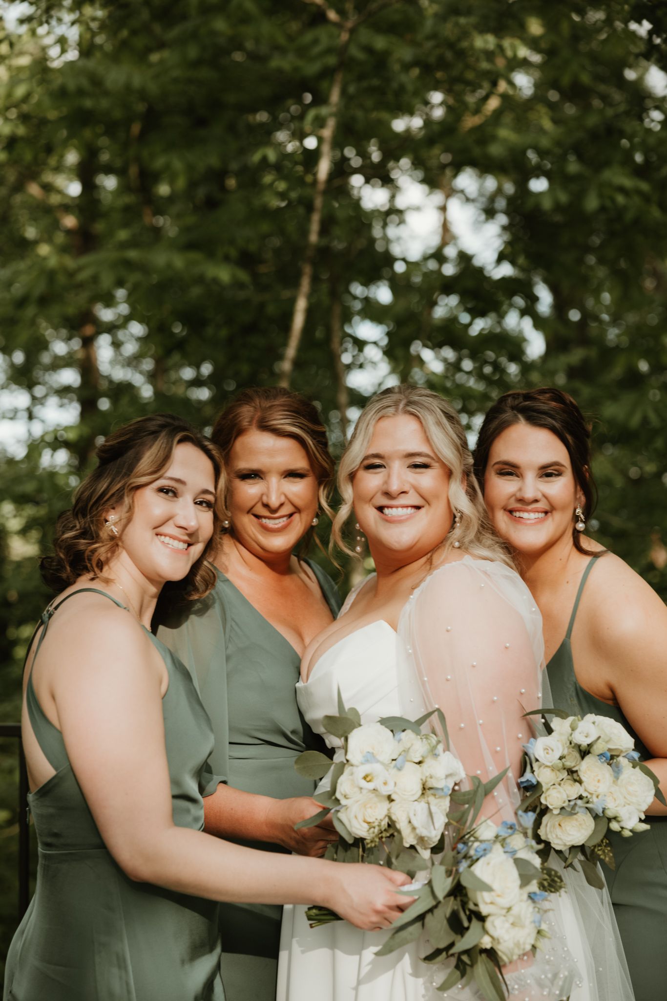 Bridal party holding white flower bouquets while wearing sage green and blush colored dresses outdoors.