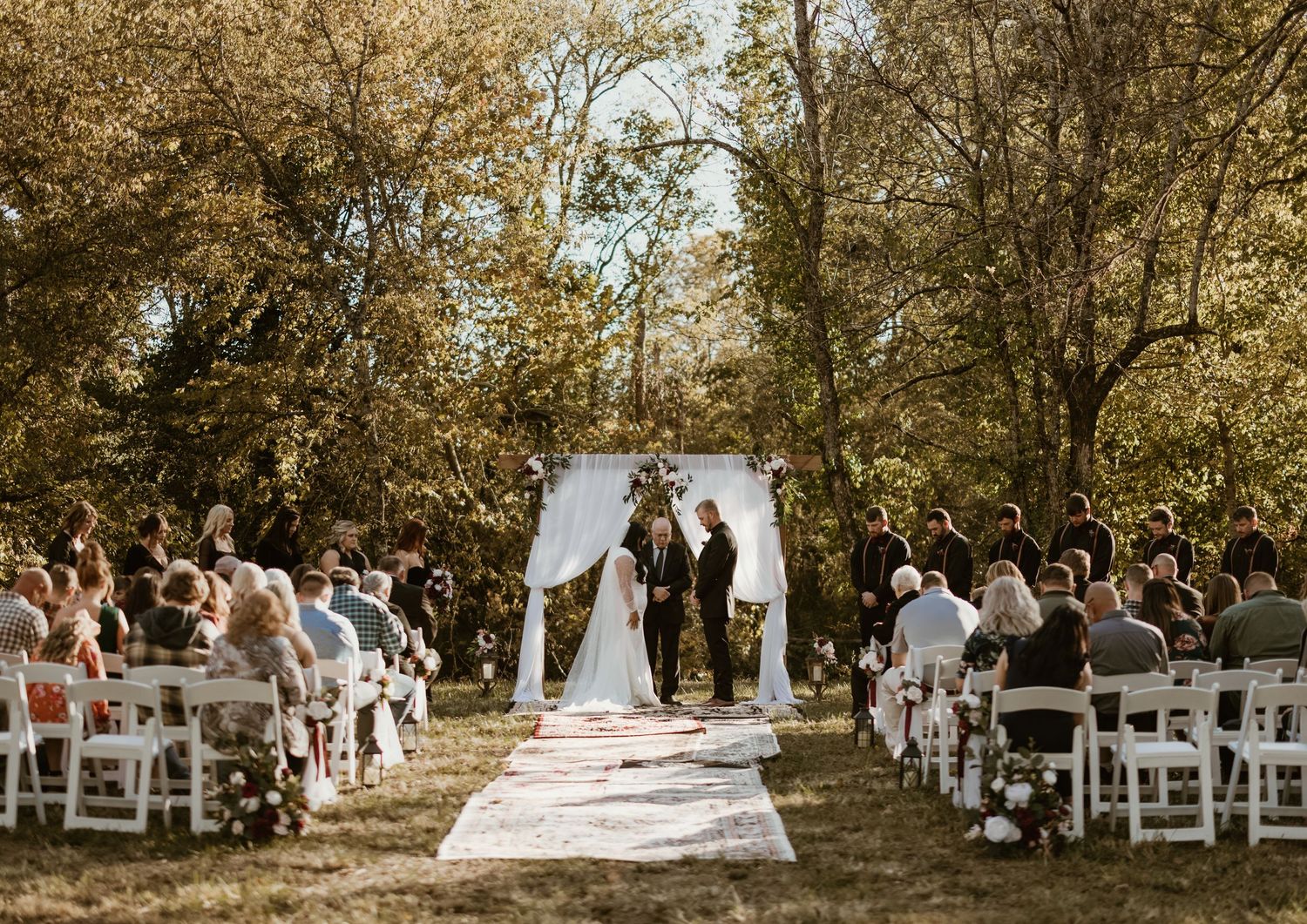 A beautiful outdoor wedding ceremony in a wooded area with white chairs and a simple white arch.