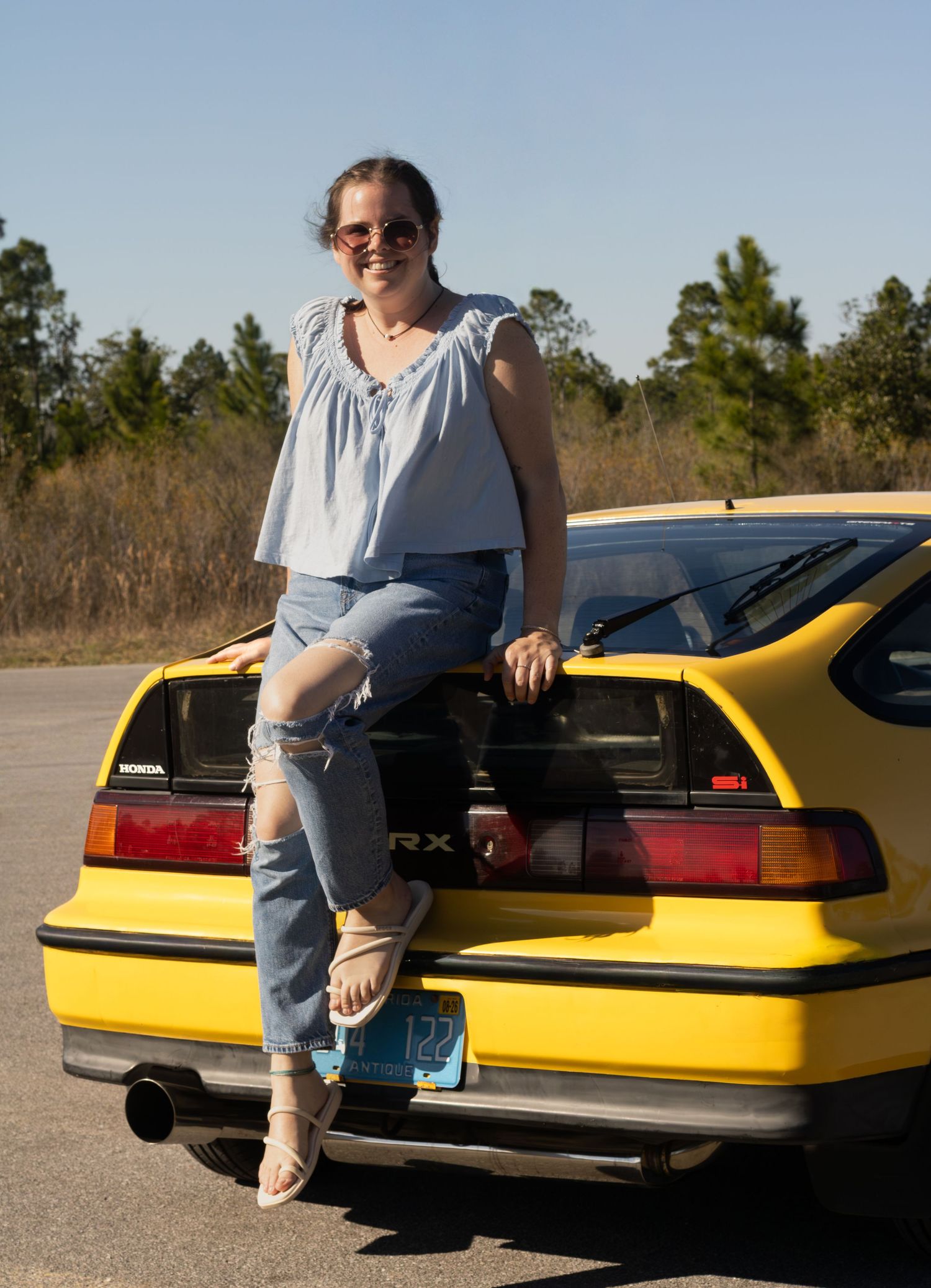 A warm picture of a women sitting on her car during summer. The picture is bright and makes her and the car pop.