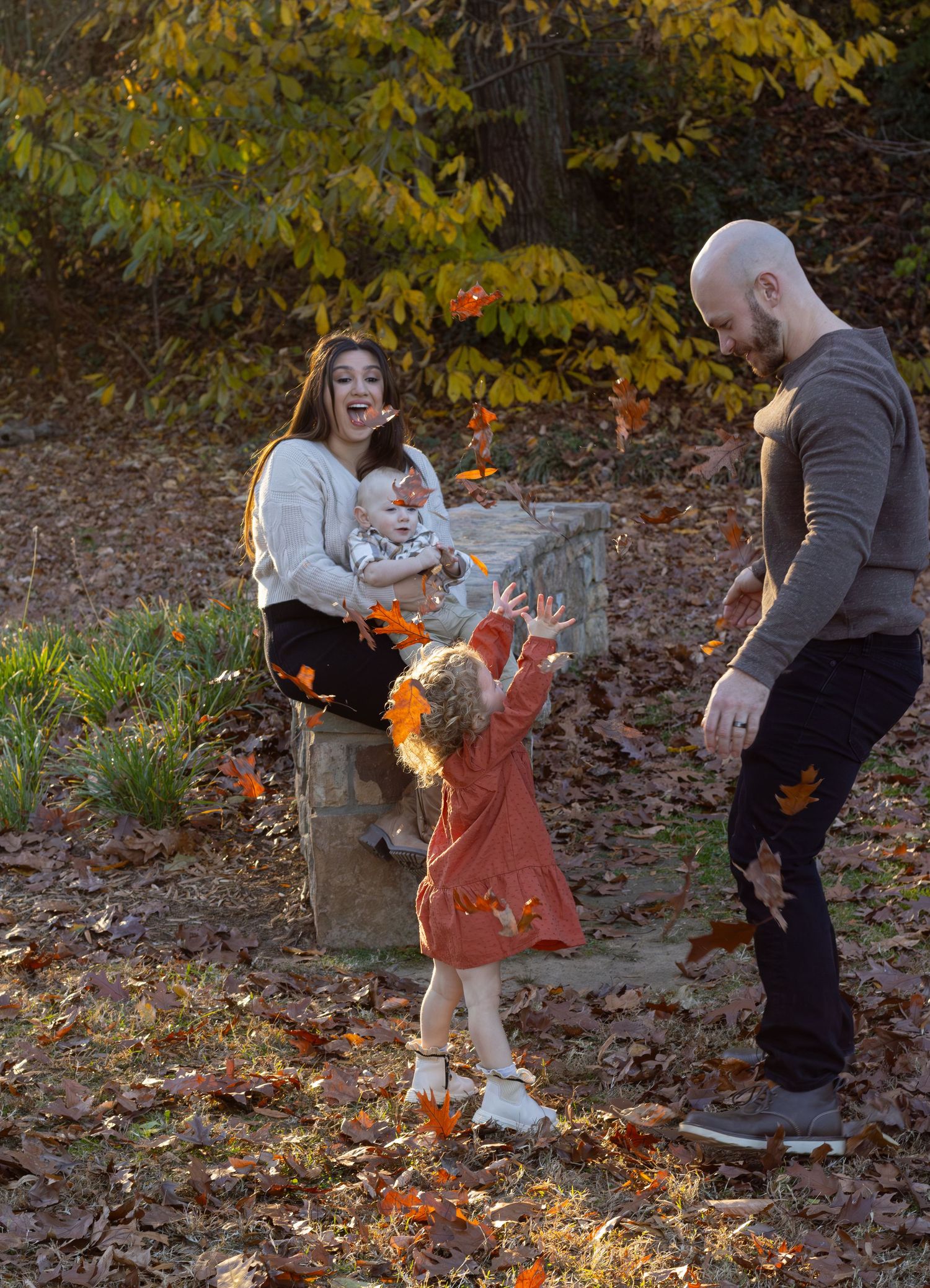 A family photo throwing up leaves in fall with the sun backlighting them. It's a family of three at a park ja