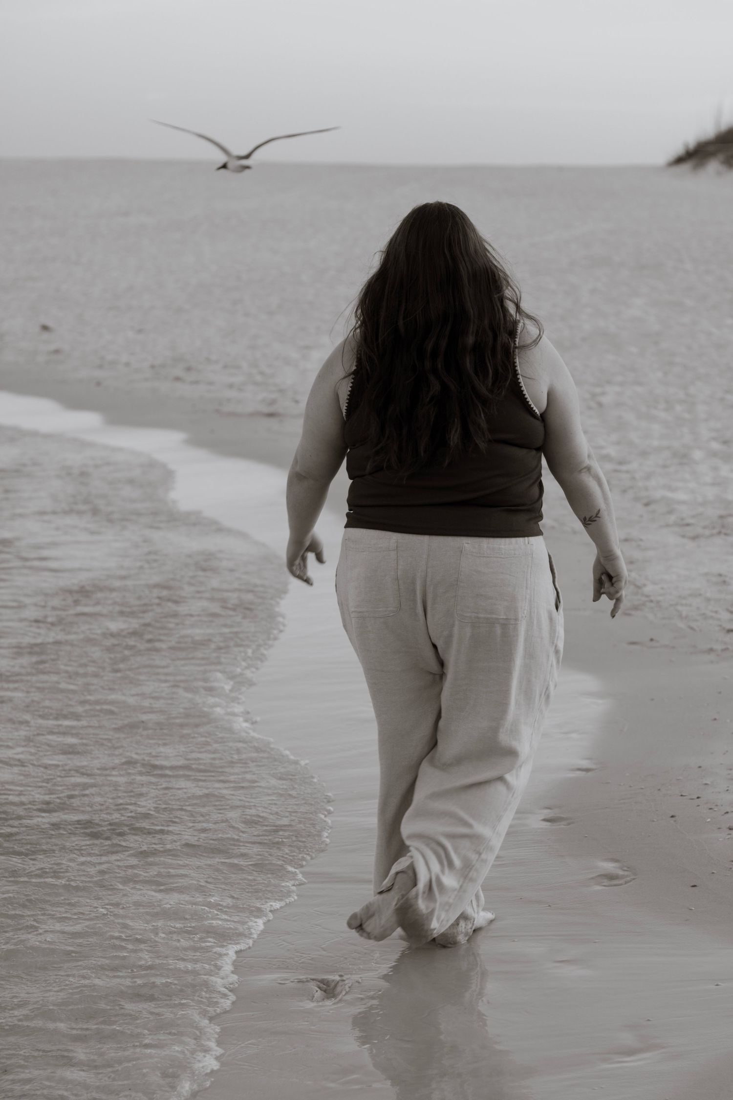A portrait of a women walking on the beach in black and white. there is a bird flying in the sky and the ocean water receding.
