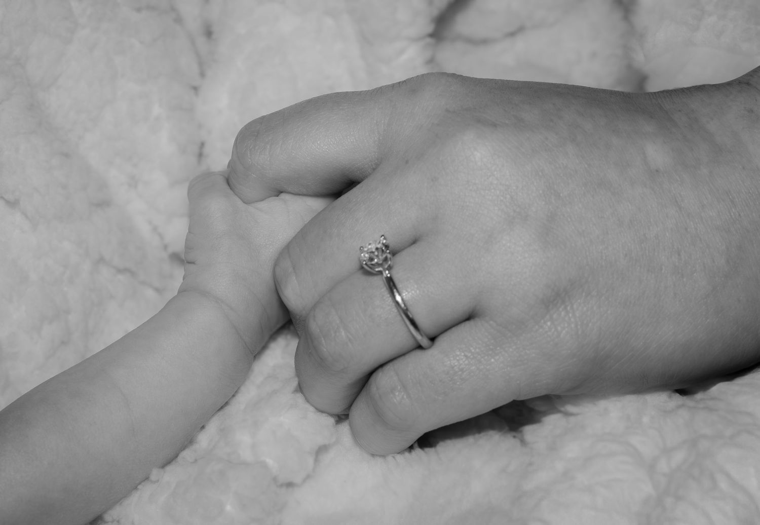 A mother and newborn son holding hands in black and white. The ring is showing and the background is fuzzy making the hands stand out.