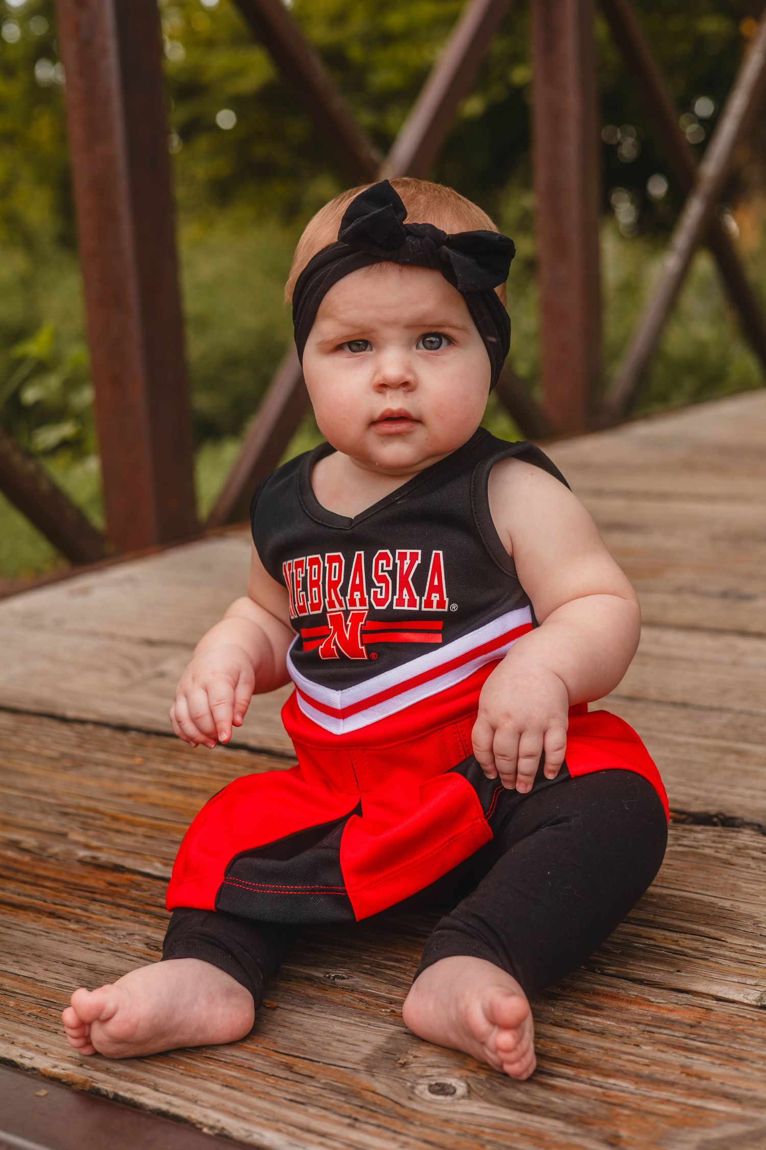Baby girl in Nebraska Cornhuskers cheerleader outfit sitting on wooden bridge in outdoor setting.