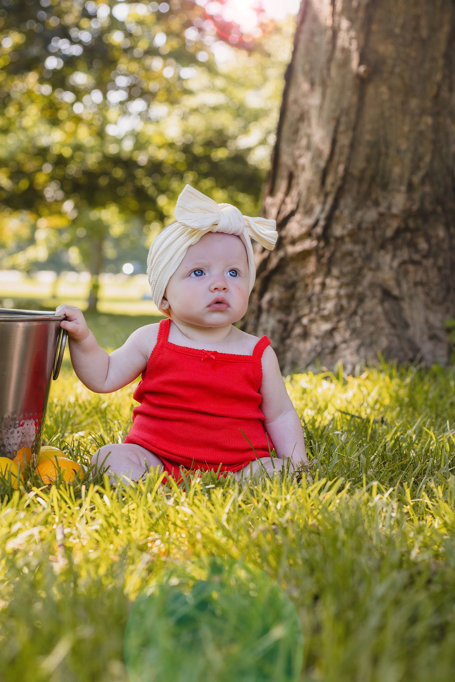 Blue-eyed baby girl in red outfit with cream bow headband sitting in sunny green grass by tree.