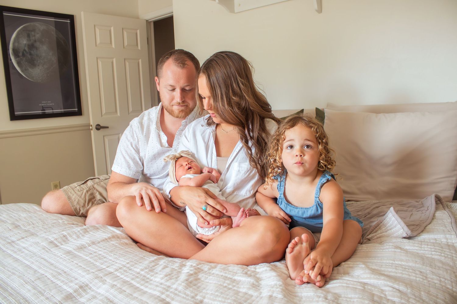 Happy family of four sitting on bed, parents admiring newborn baby while toddler sits beside them.
