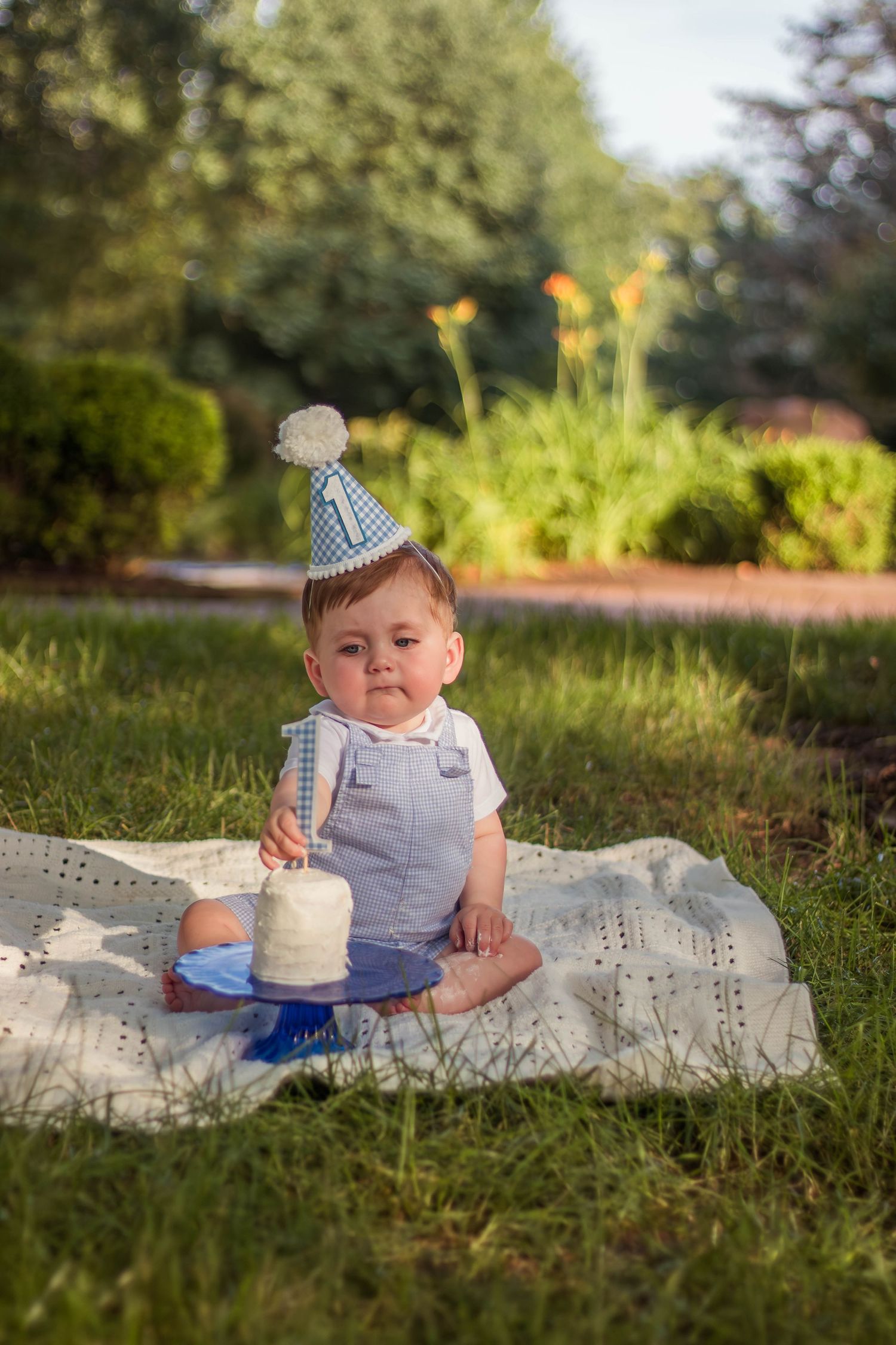 Baby in blue outfit and party hat sitting on blanket outdoors with first birthday smash cake.