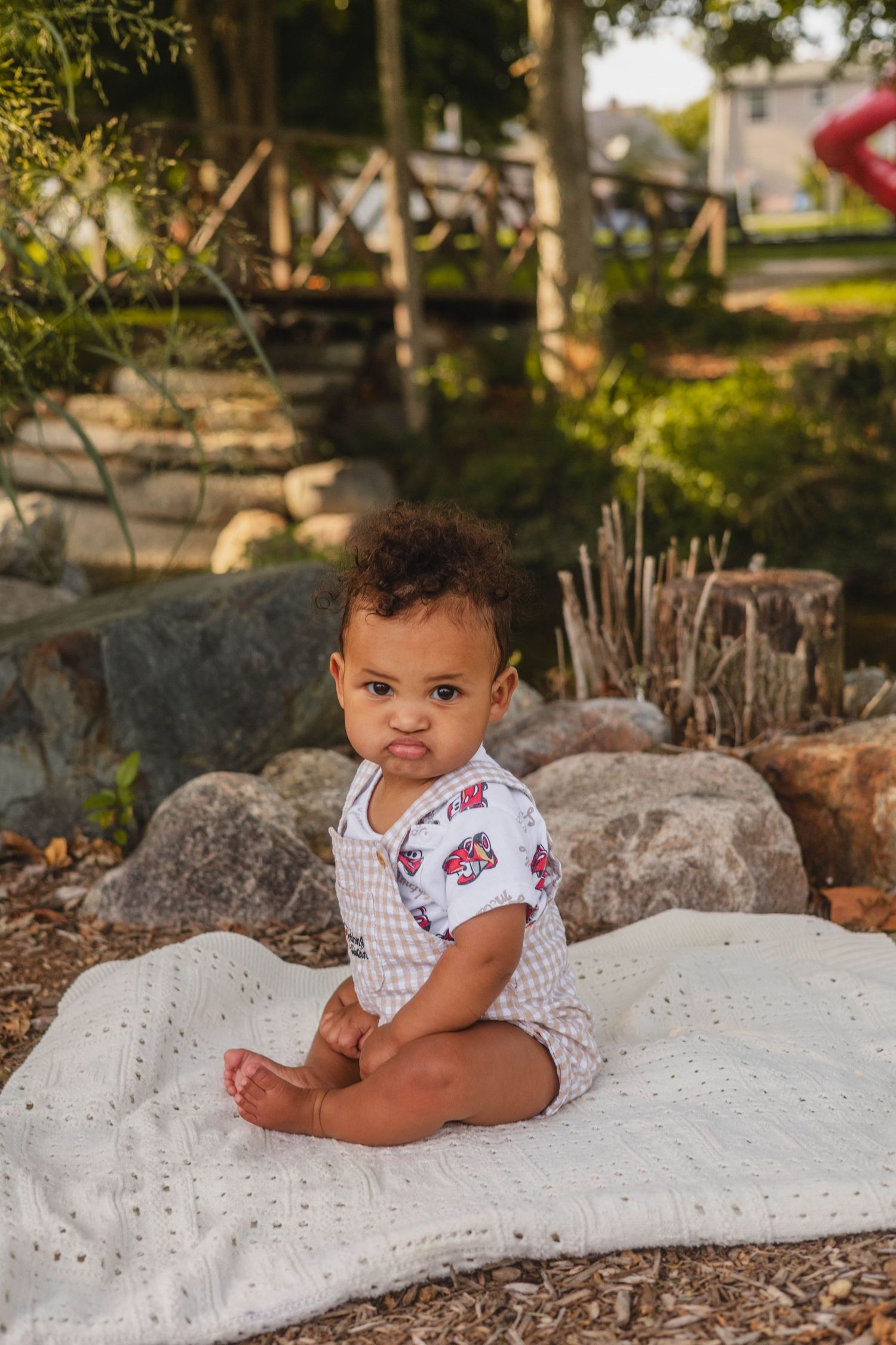 Adorable baby sitting on white blanket outdoors on rocks in printed onesie, making a cute expression.