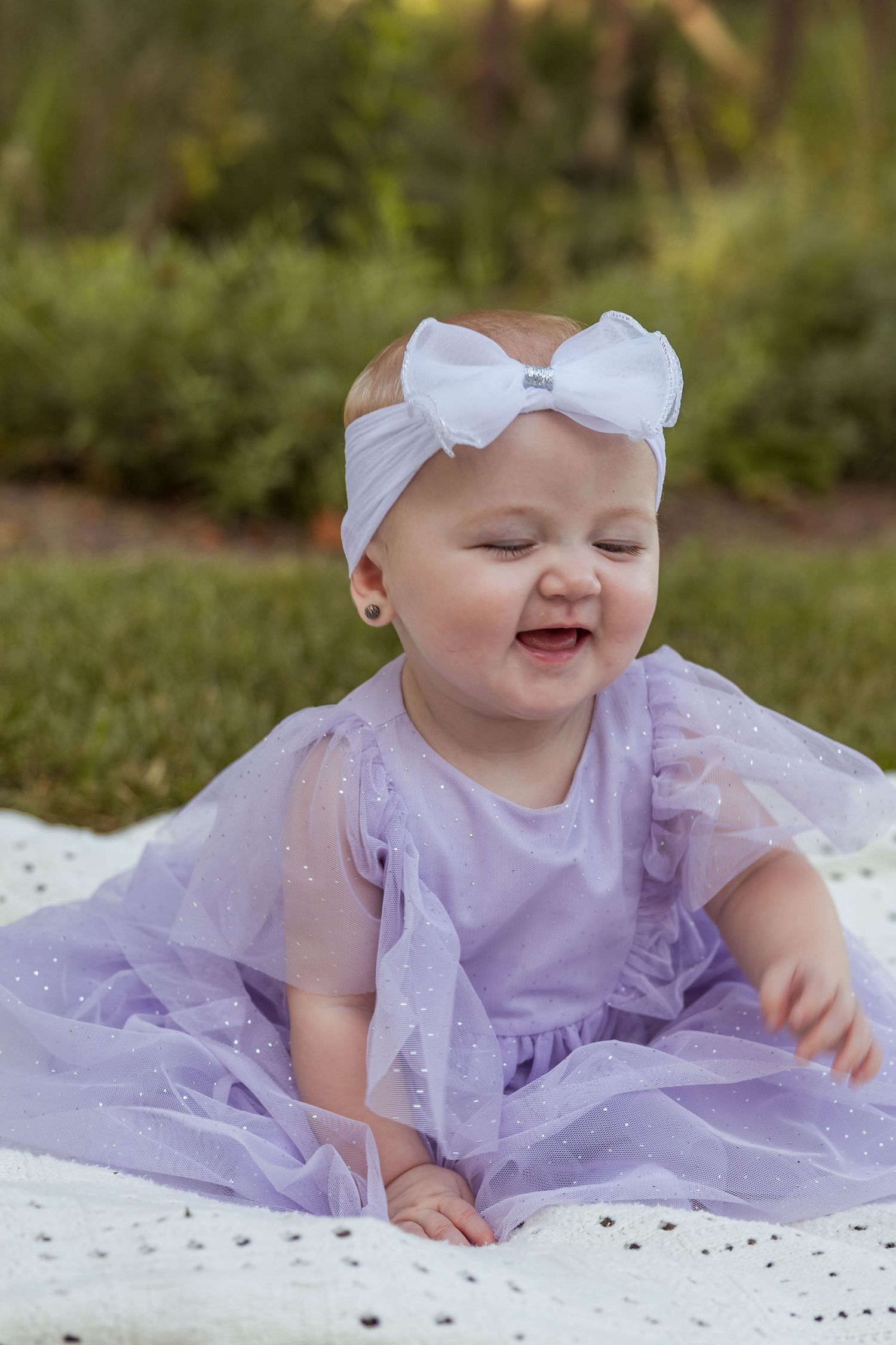 Joyful baby girl laughing in sparkly lavender tulle dress with white bow headband outdoors.