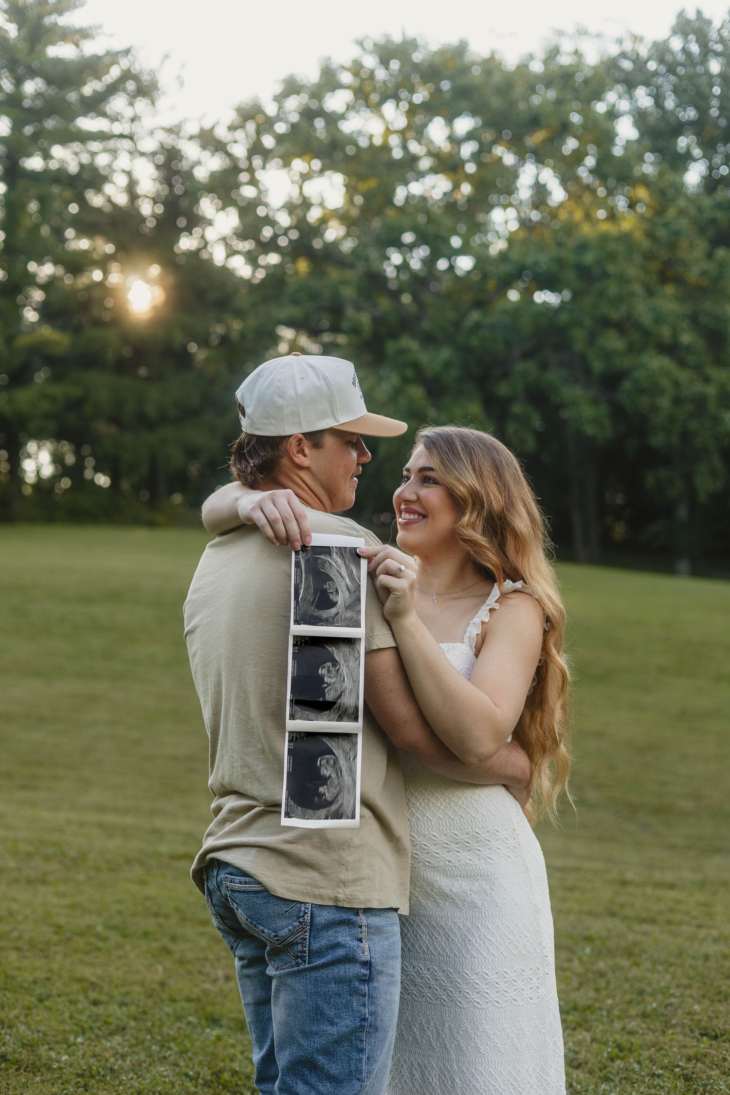 Happy couple holding ultrasound photos embracing in park at sunset during maternity photography session.