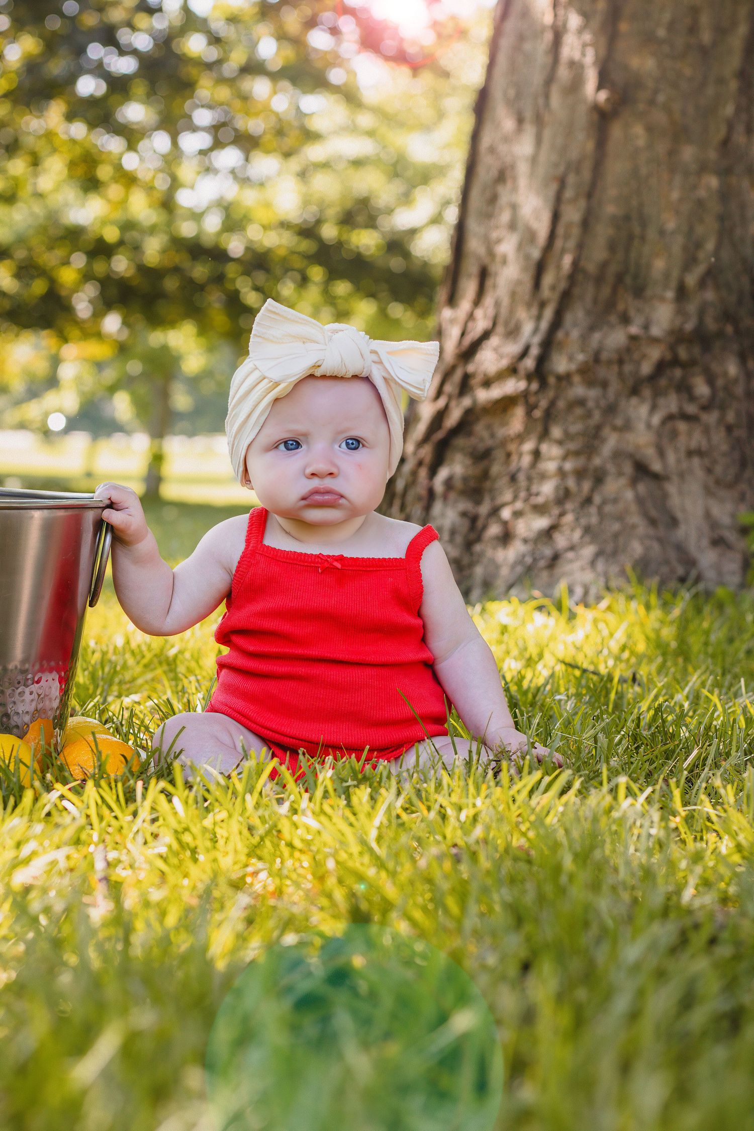 Cute baby girl in red romper with cream bow headband sitting in grass by tree in sunny park.