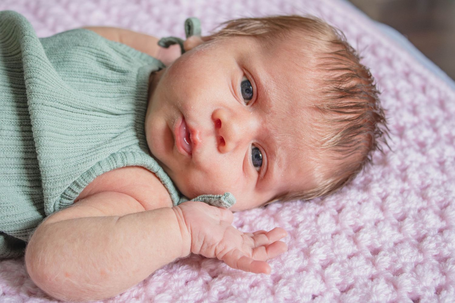 Newborn baby in green outfit lying on pink crochet blanket with eyes open.