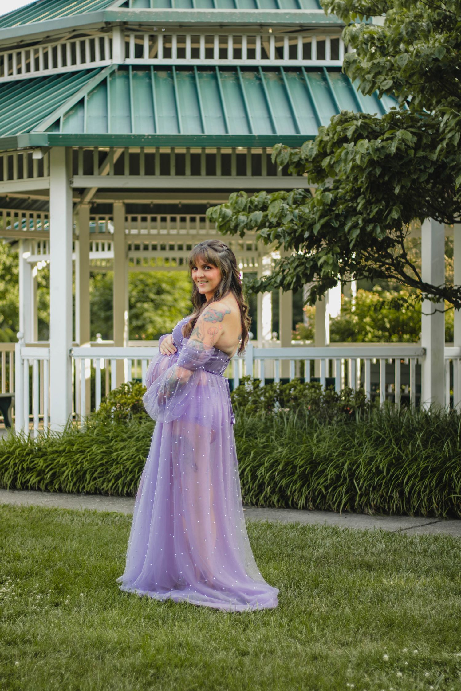 Smiling pregnant woman in purple tulle gown posing near a white gazebo outdoors.
