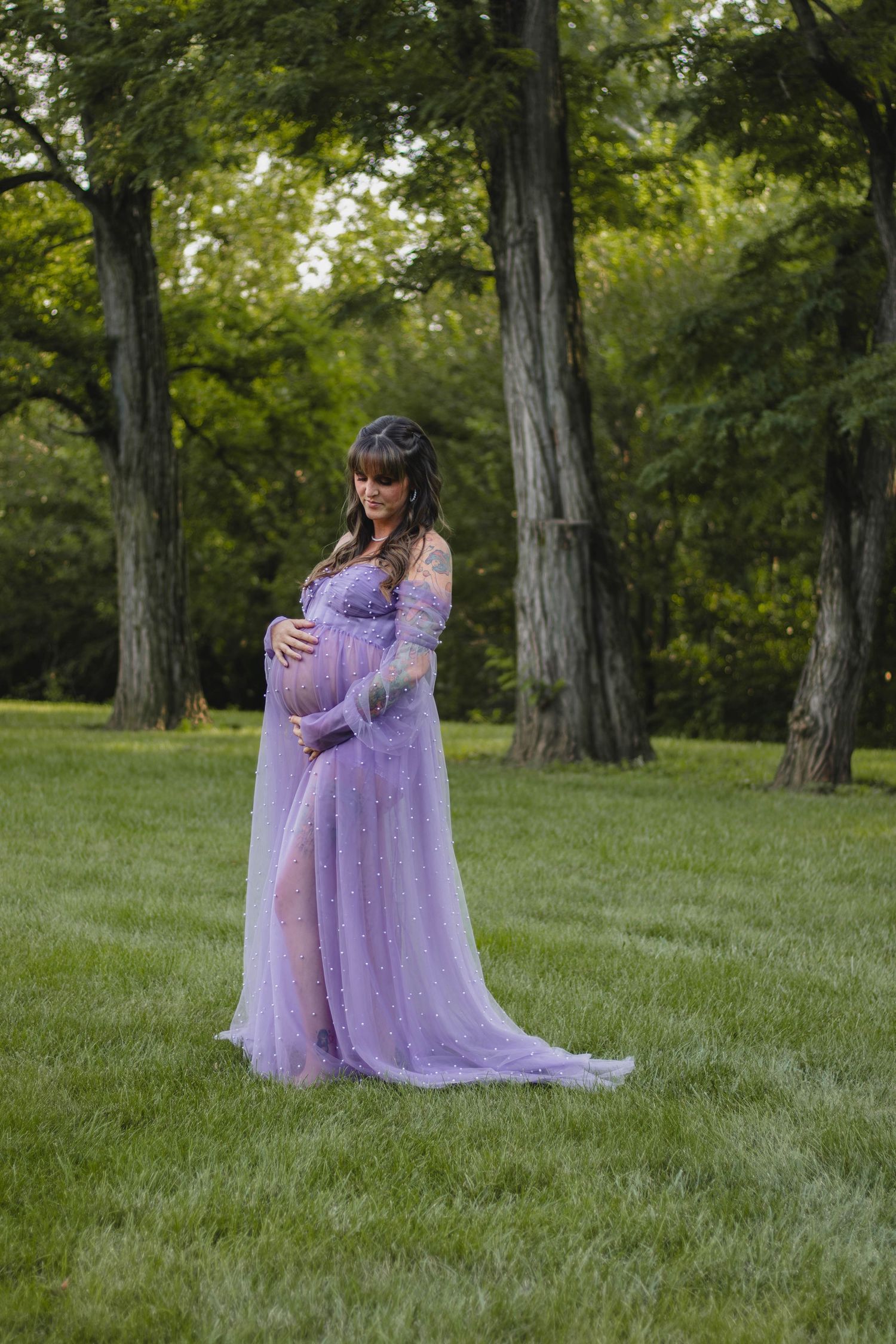 Pregnant woman in flowing lavender tulle gown cradling belly while standing in lush green park.