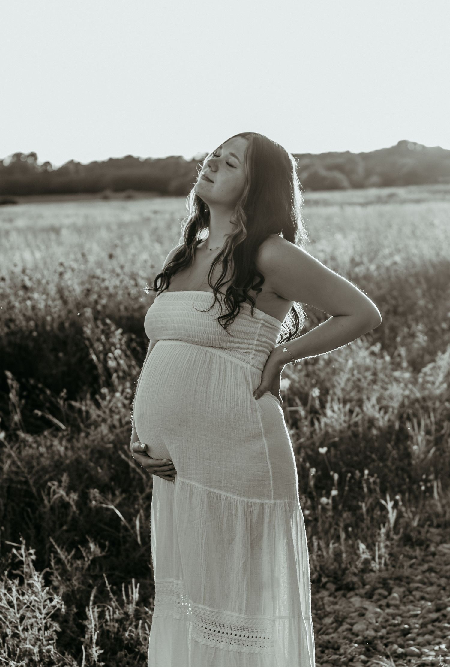 Black and white photo of pregnant woman in white dress standing in a sunlit meadow.