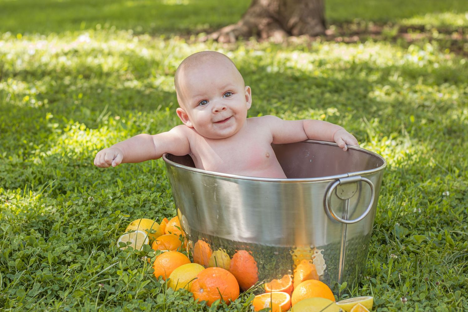 Smiling baby sitting in a silver metal tub outdoors surrounded by colorful pumpkins and oranges.