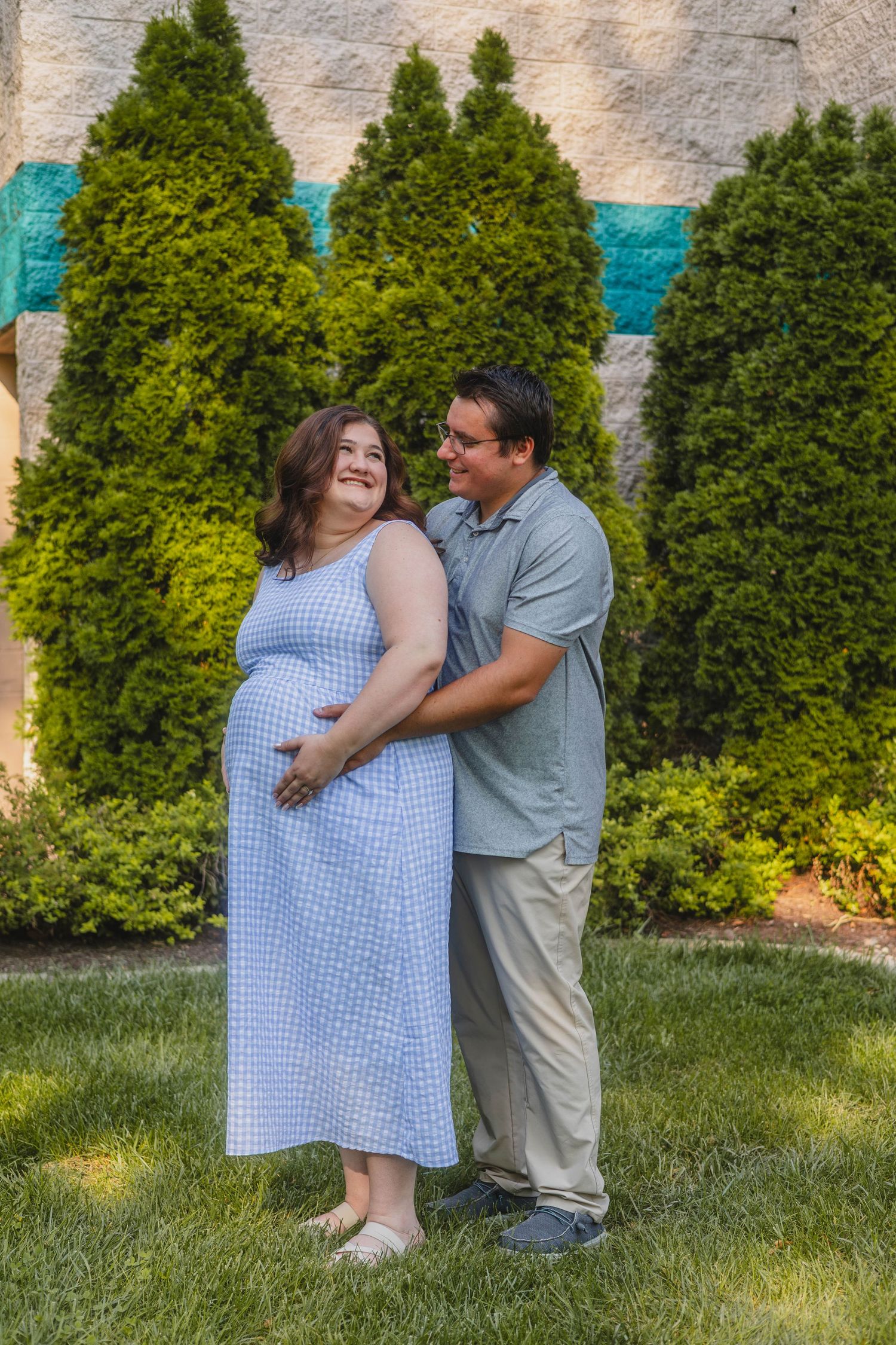 Couple embracing outdoors with man holding pregnant womans belly in blue gingham dress near evergreens.