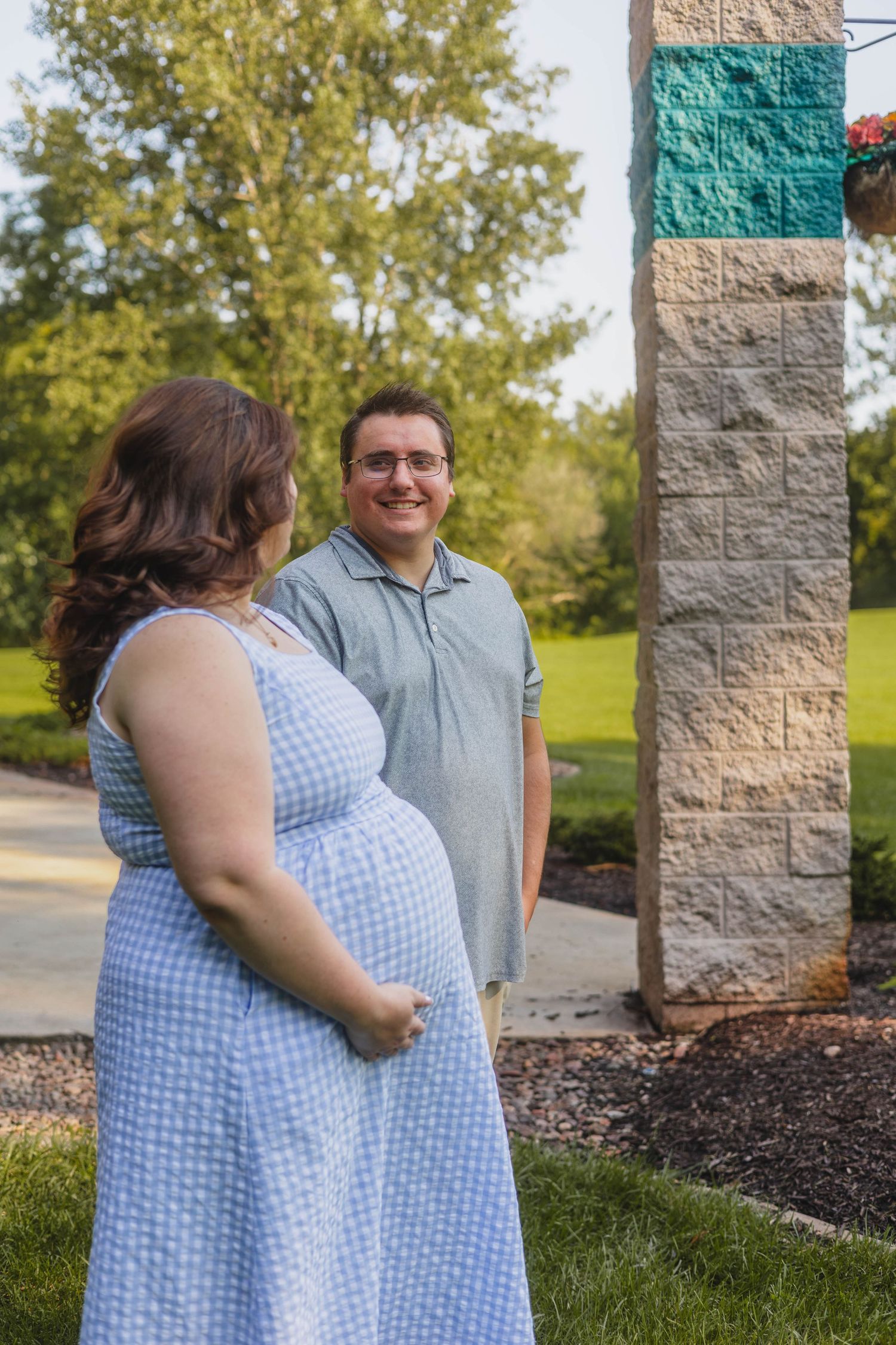 Pregnant woman in blue gingham dress with partner smiling outdoors near stone column.