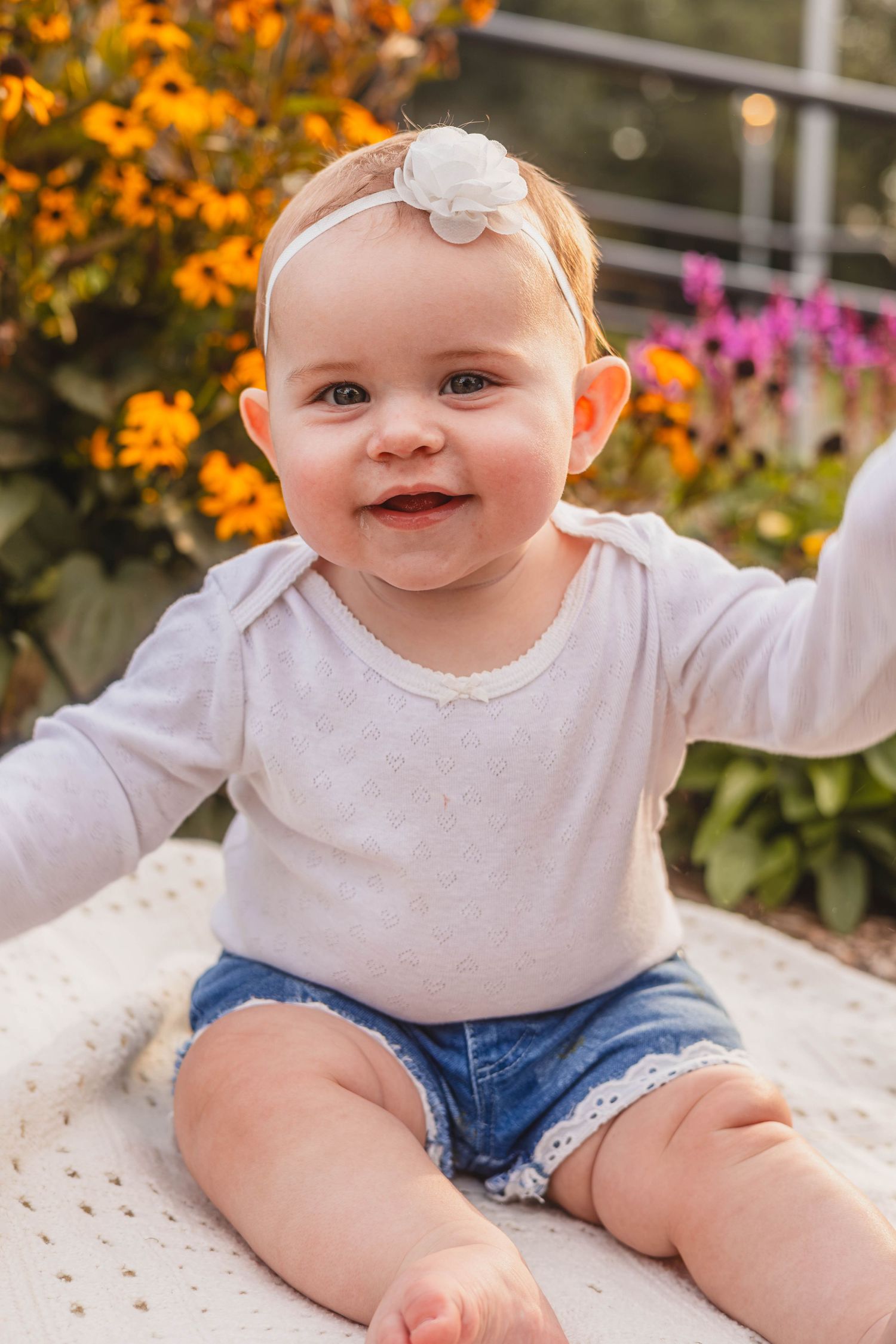 Happy smiling baby girl in white bodysuit and denim shorts with floral headband sitting outdoors.