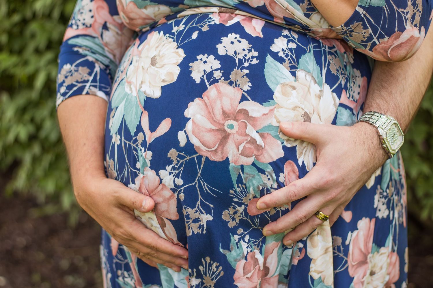 Close-up of pregnant belly in blue floral dress with partner's hands gently cradling bump.