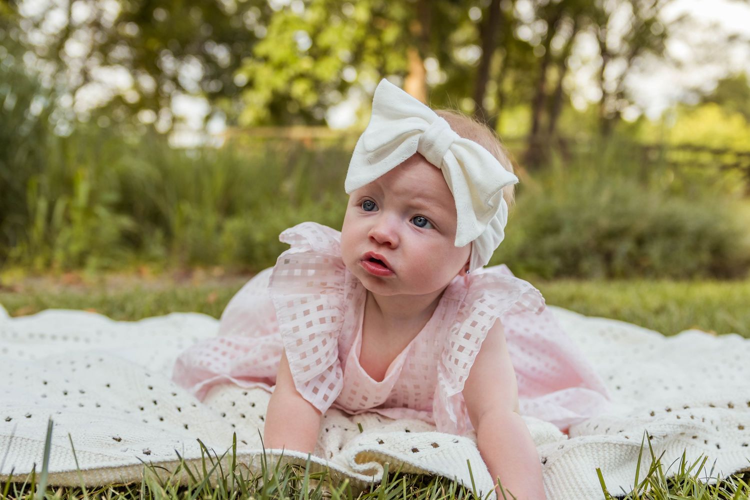 Sweet baby girl in pink dress and white bow headband lying on white blanket in green park.