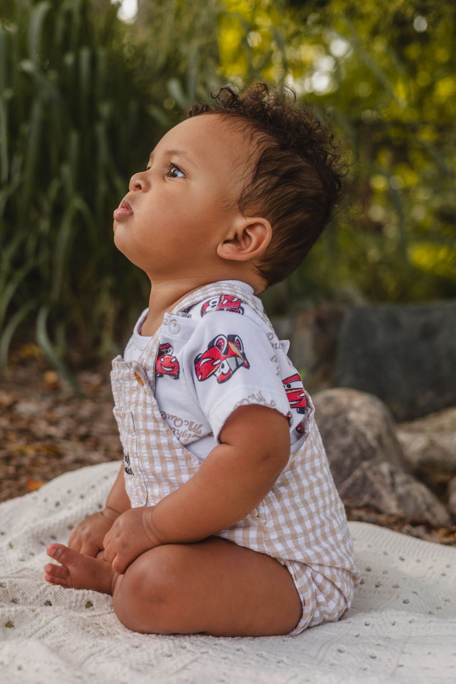 Cute baby boy in gingham overalls sitting on white blanket looking up outdoors.