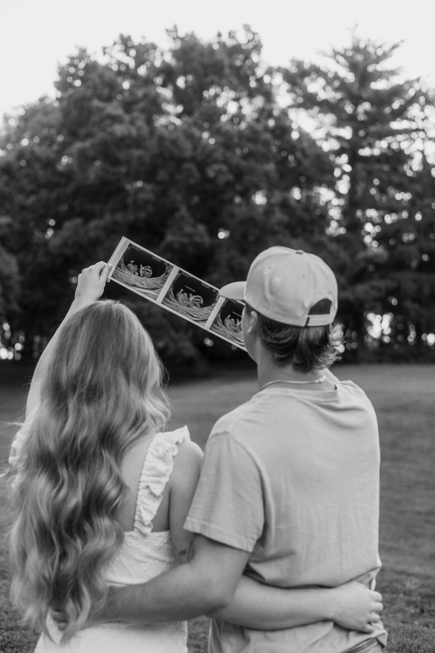 Black and white photo of couple holding ultrasound images together in park setting.